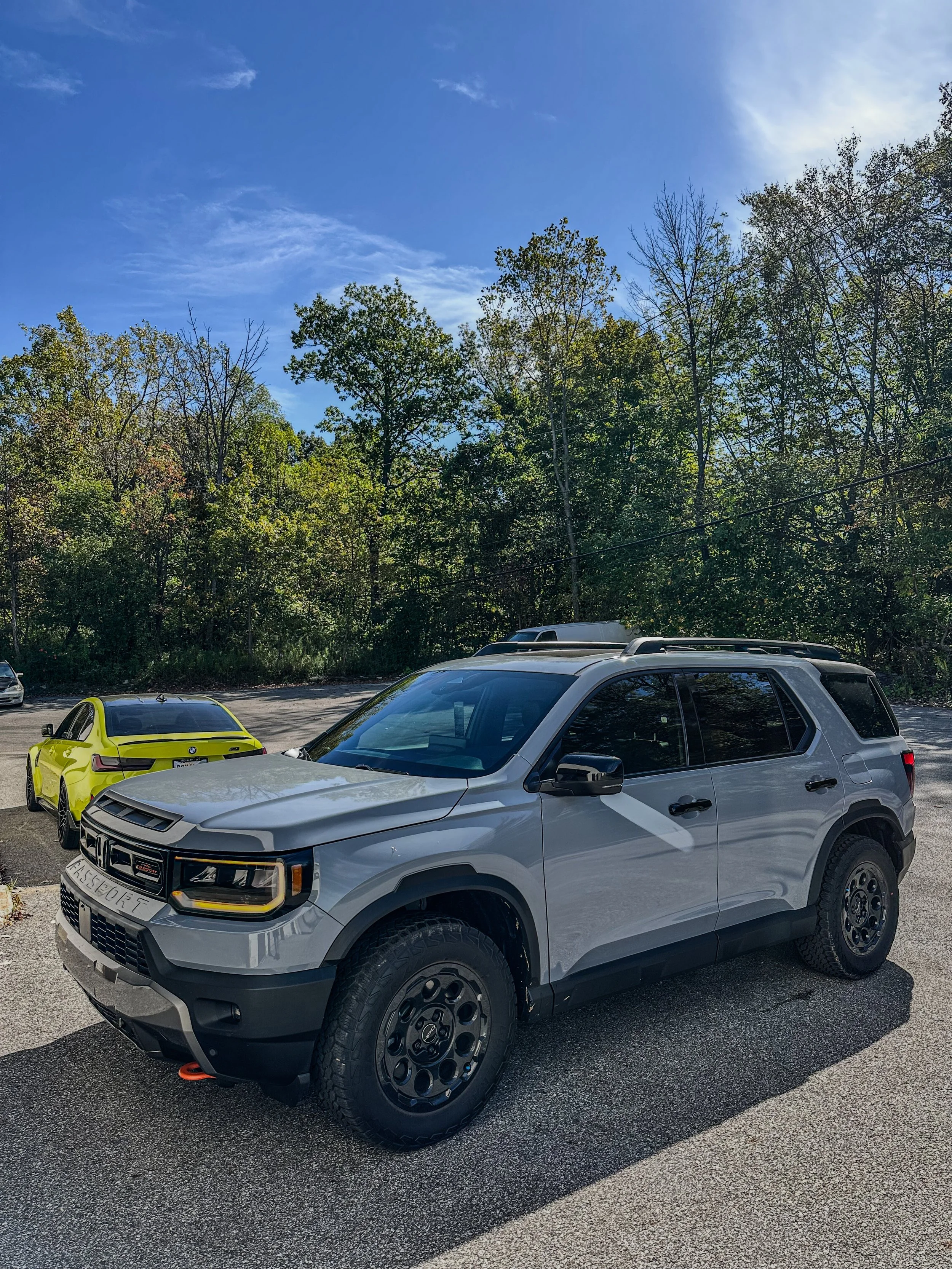 A silver compact SUV parked outdoors on a paved lot with a yellow sports car and other vehicles in the background. Trees and a blue sky with some clouds are seen behind the vehicles.