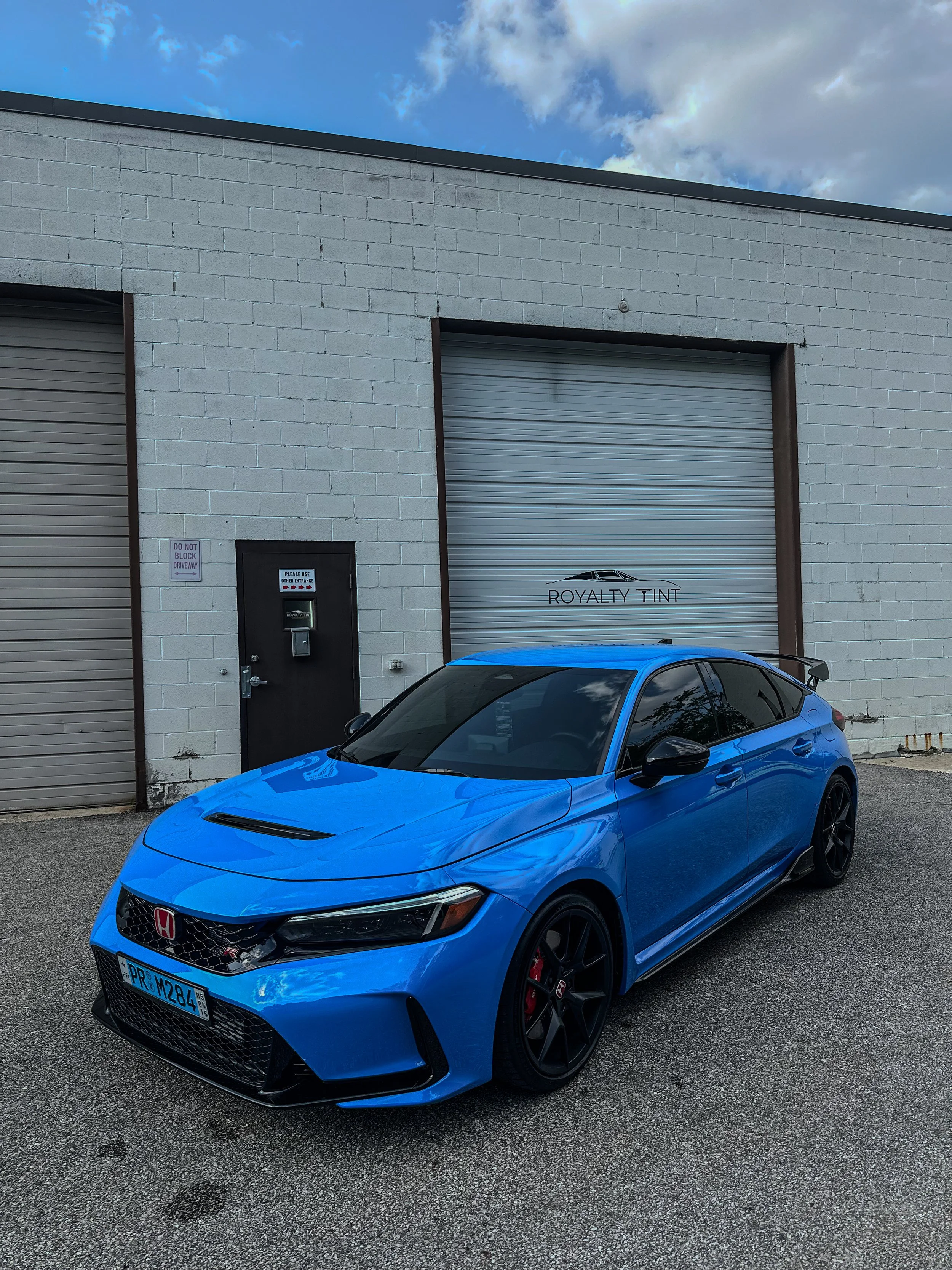 A blue Honda Civic Type R parked in front of a white brick building with rolling garage doors and signage that says Royalty Tint.