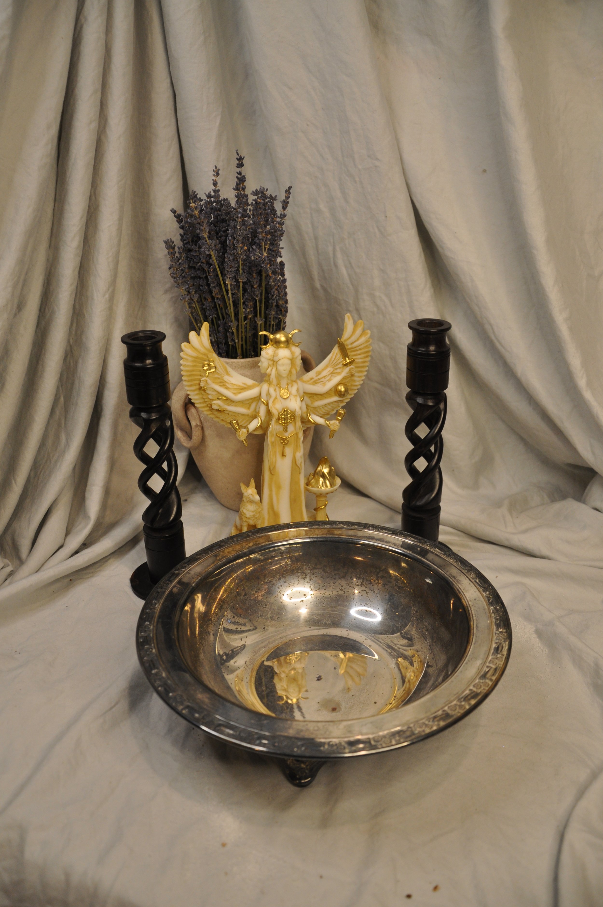 A decorative setup with a brass statue of a multi-armed deity, a vase of dried lavender, black candle holders, and a silver bowl on a draped beige fabric background.