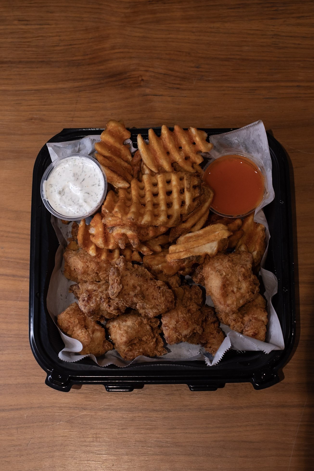 Black tray on wooden table. Tray contains waffle fries and nuggets. Two ramequins filled with sauce.