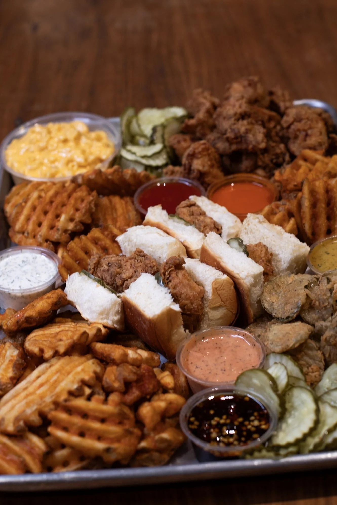 A tray with a container filled with Mac and cheese, pickle slices, chicken nuggets, waffle fries, chicken sliders, and fried pickles, as well as ramequins with sauce.