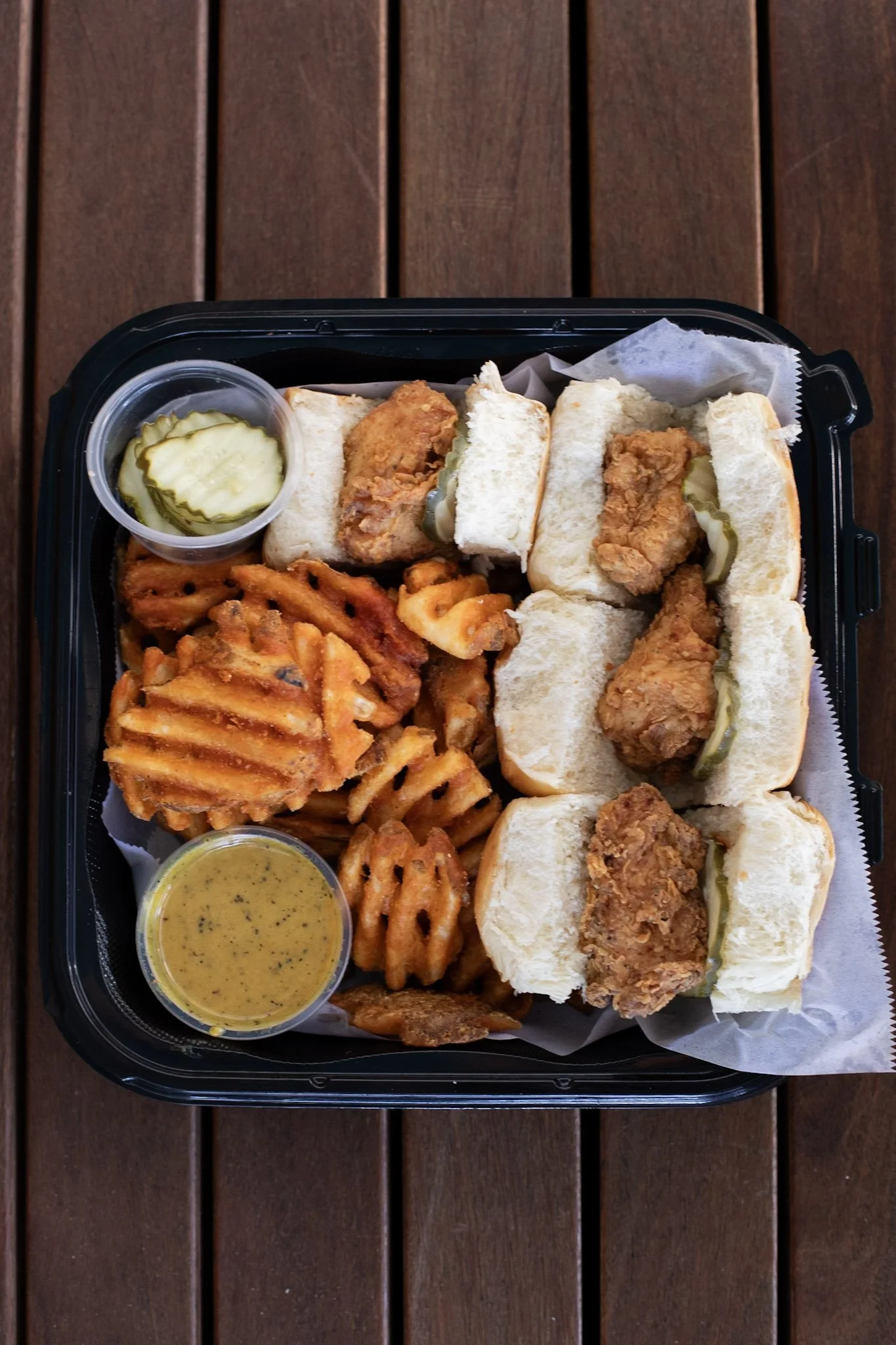 Black Tray on wooden table, black tray is filled with chicken sliders, waffle fries, two ramequins, one with pickles, one with sauce.