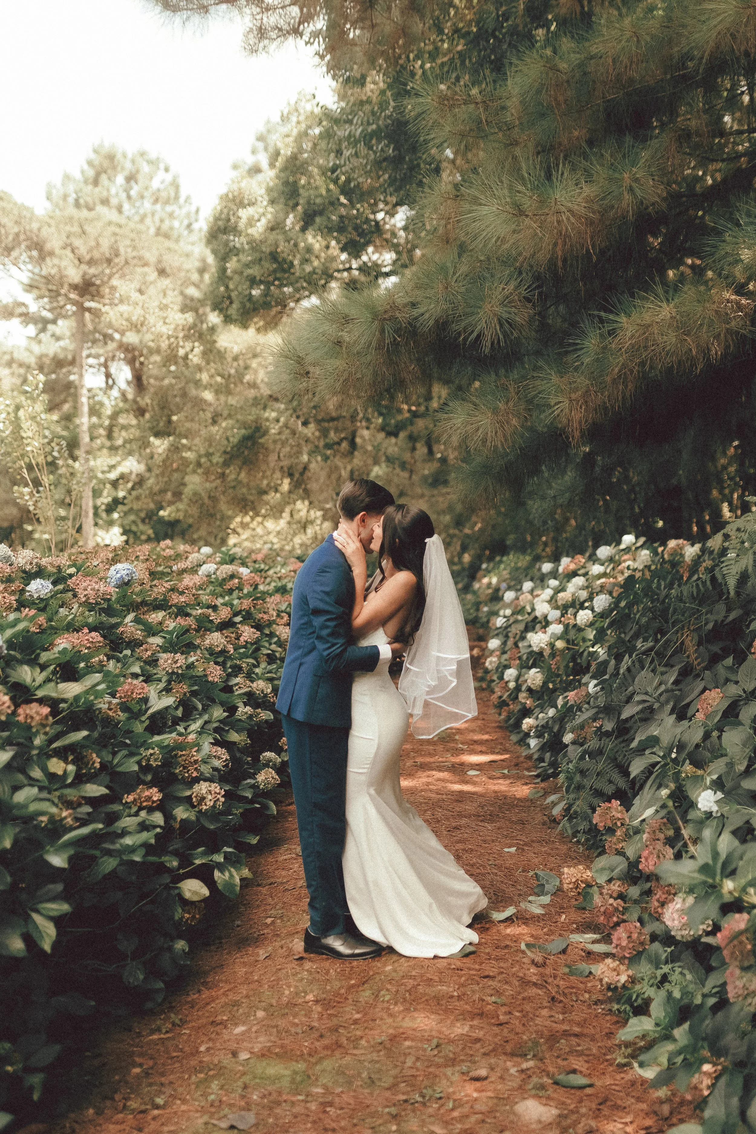 A bride and groom sharing a kiss on a forested wedding path, surrounded by flowering bushes and tall trees.