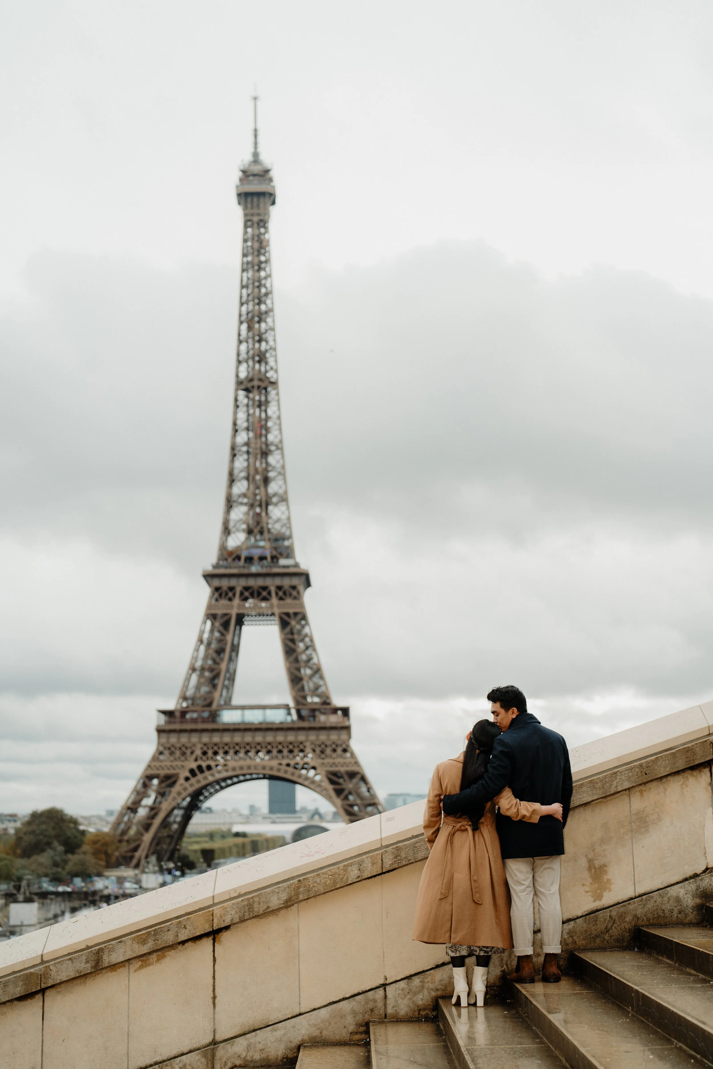 A couple embraces on a staircase near the Eiffel Tower in Paris, under a cloudy sky.