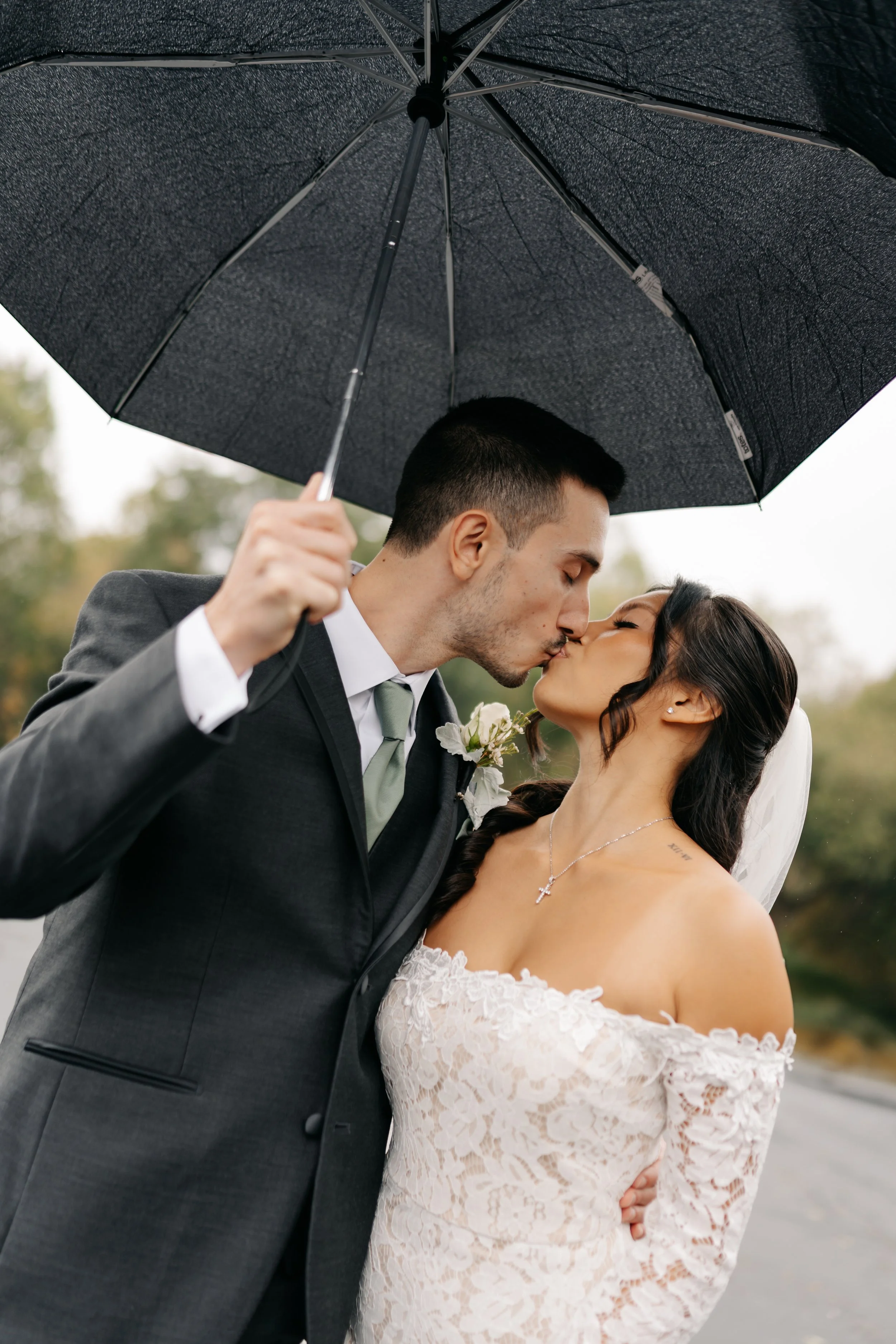 A wedding couple, a man in a suit and a woman in a lace dress, sharing a kiss under a black umbrella outdoors on a rainy day.
