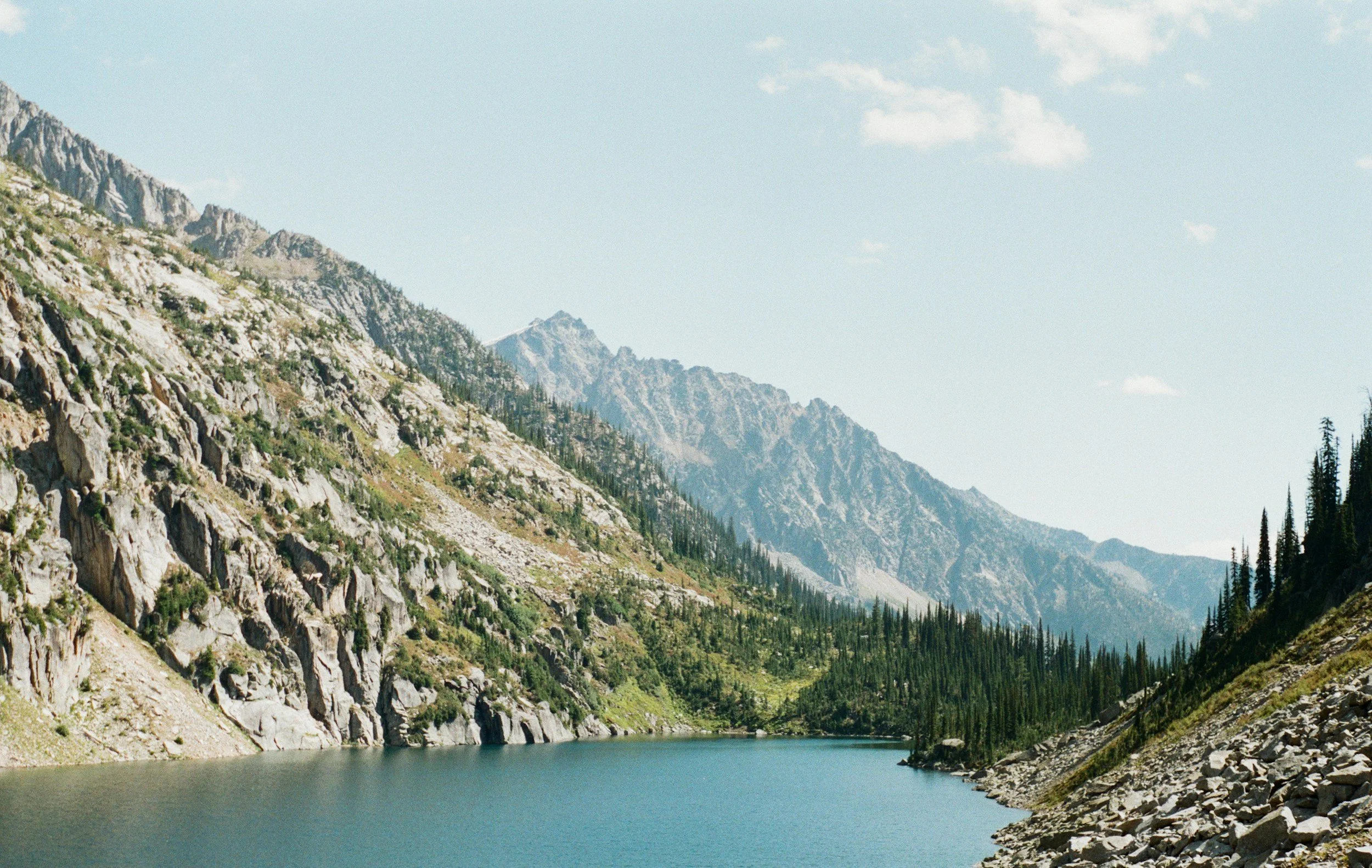 Scenic view of a mountain lake surrounded by rocky slopes and pine trees, with mountains in the background under a partly cloudy sky.