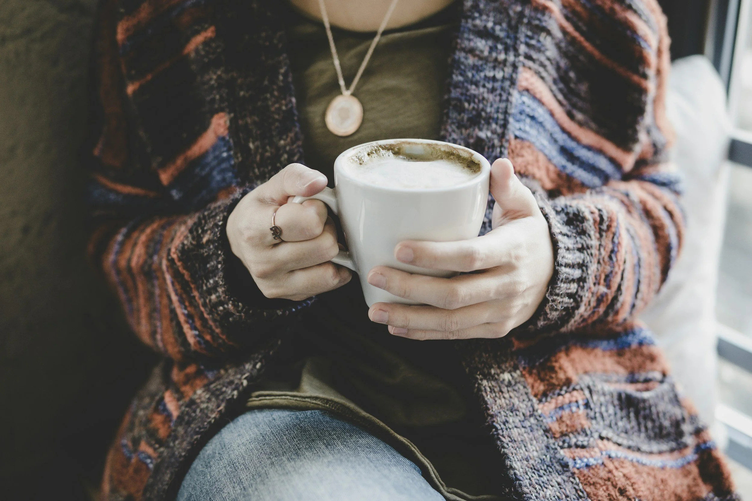Person wearing a multicolored striped sweater, holding a white mug of coffee or hot chocolate, sitting by a window.
