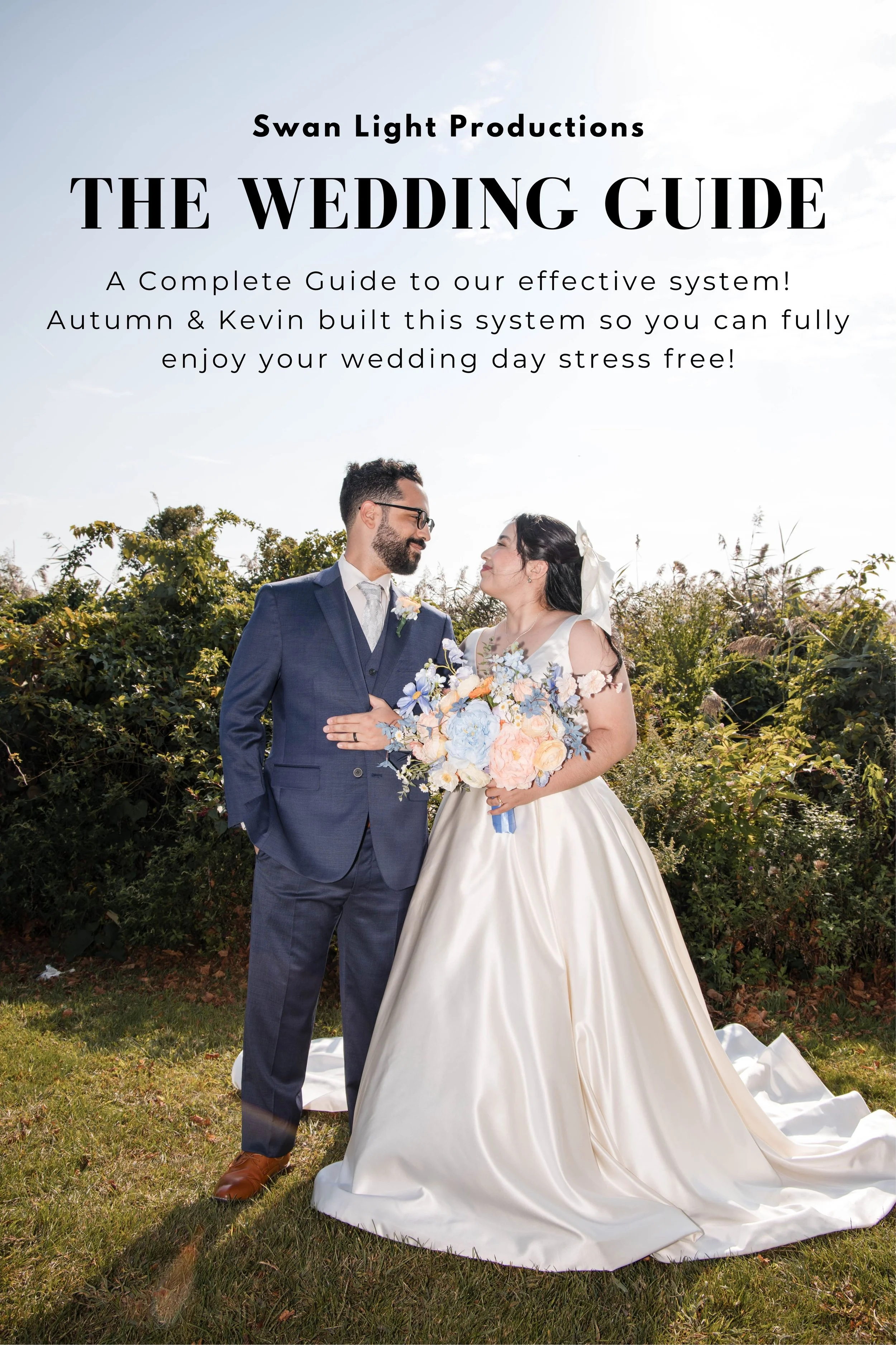 A bride and groom standing outdoors on grass in front of bushes, looking at each other. The bride is wearing a white wedding gown and holding a bouquet, while the groom is dressed in a blue suit with a tie and glasses. The sun is shining, creating a bright atmosphere.