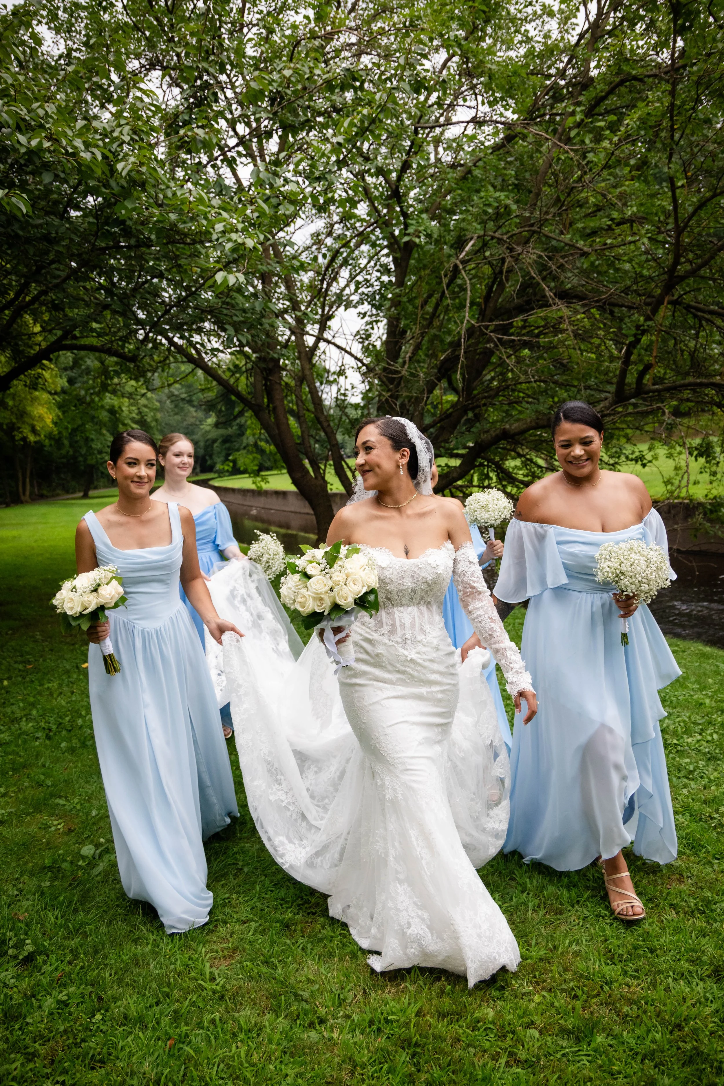 A bride in a white lace wedding gown holding a bouquet, walking with four bridesmaids in light blue dresses, in a park with green grass and trees.