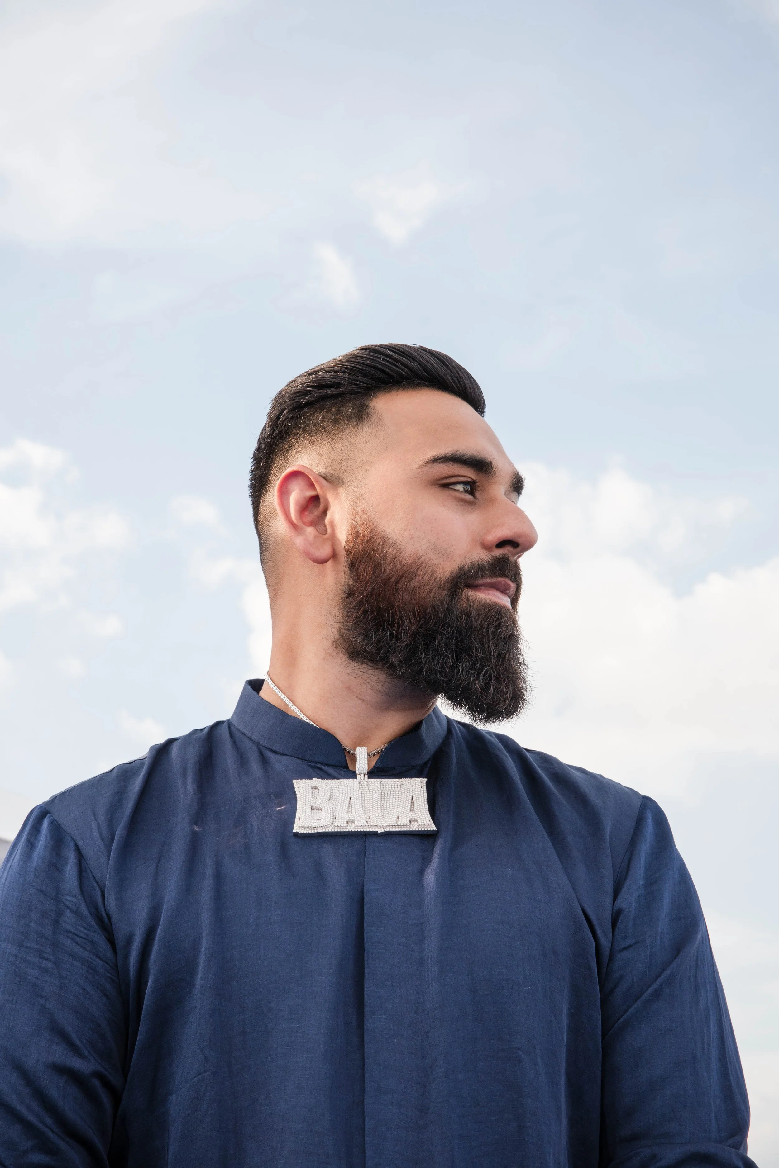 Profile of a man with a beard and styled hair wearing a blue shirt against a cloudy sky background.