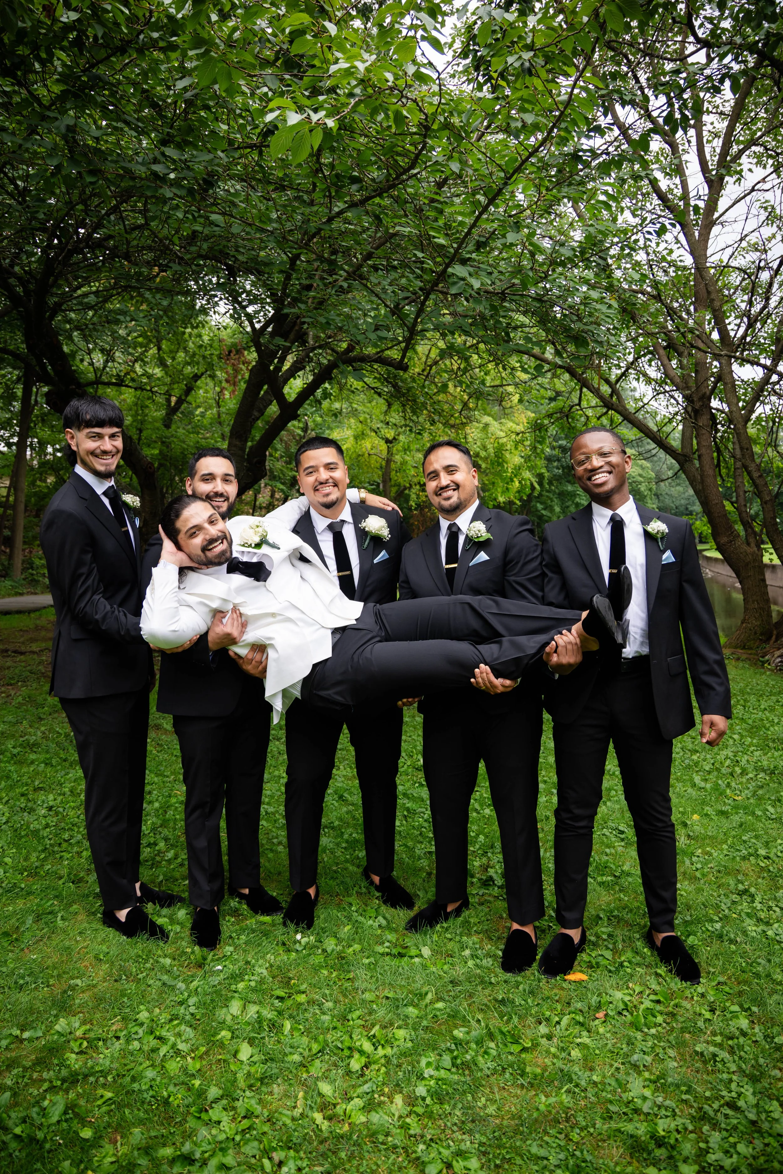 Group of six men dressed in formal black suits, white shirts, and black shoes outdoors on green grass, with some men wearing boutonnières, and one man being carried horizontally by the others, in front of trees.
