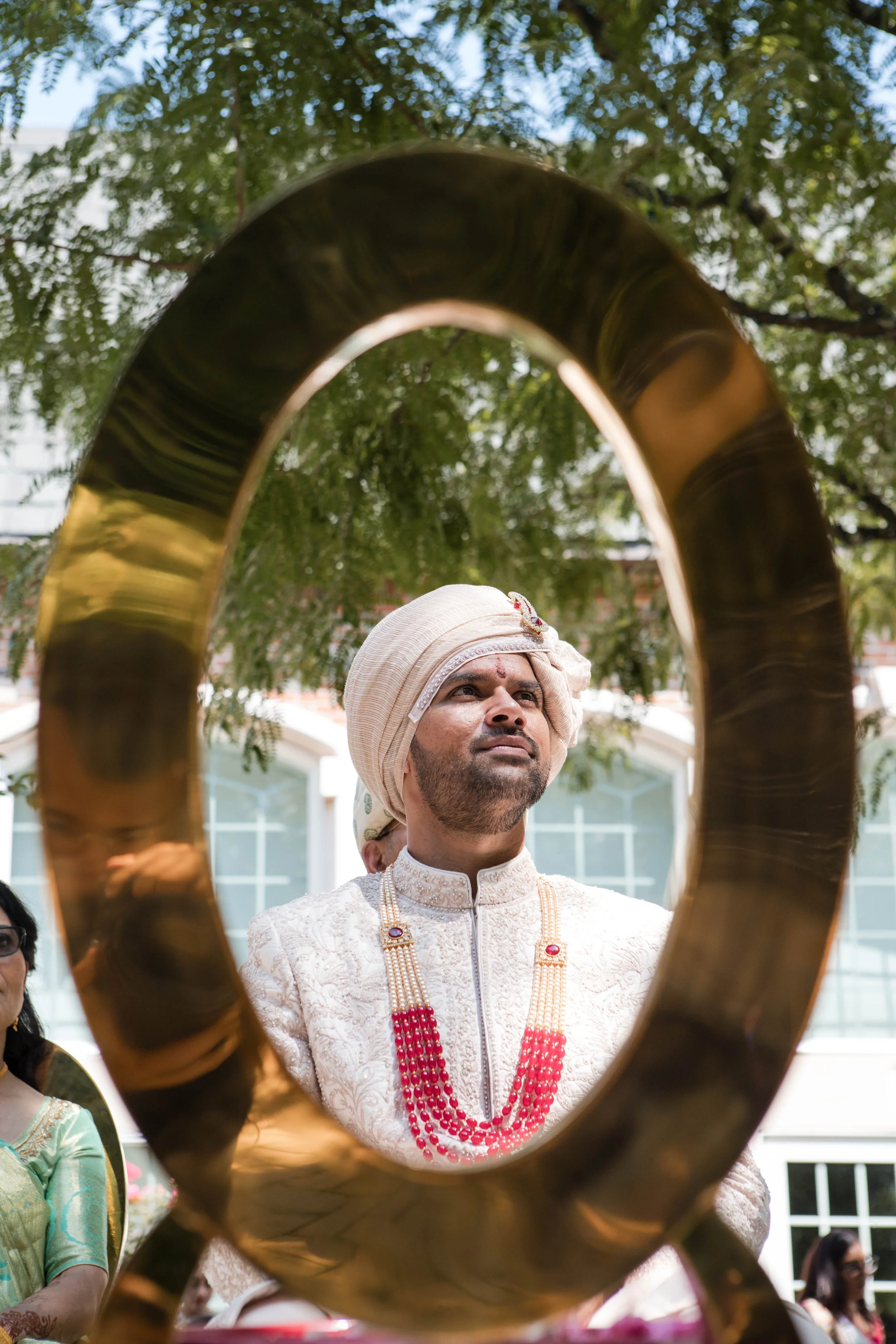 A man dressed in traditional Indian attire looking through a large brass ring at a cultural or religious event, with trees and a building in the background.