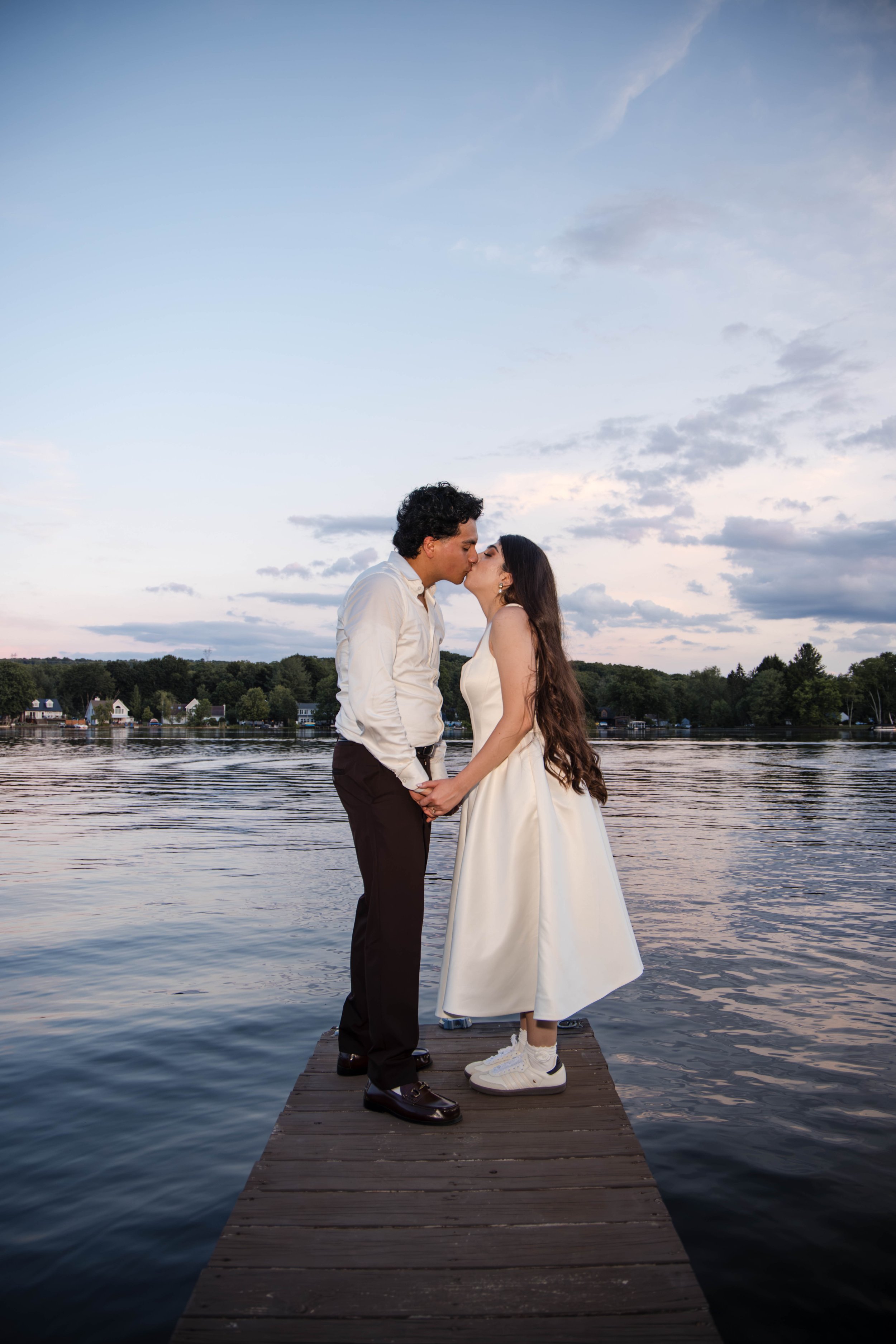 A couple stands on a wooden dock by a lake at sunset, kissing. The man wears a white shirt and dark pants, the woman wears a white dress and sneakers. The sky is cloudy with a soft sunset glow, and there are houses and trees across the lake.