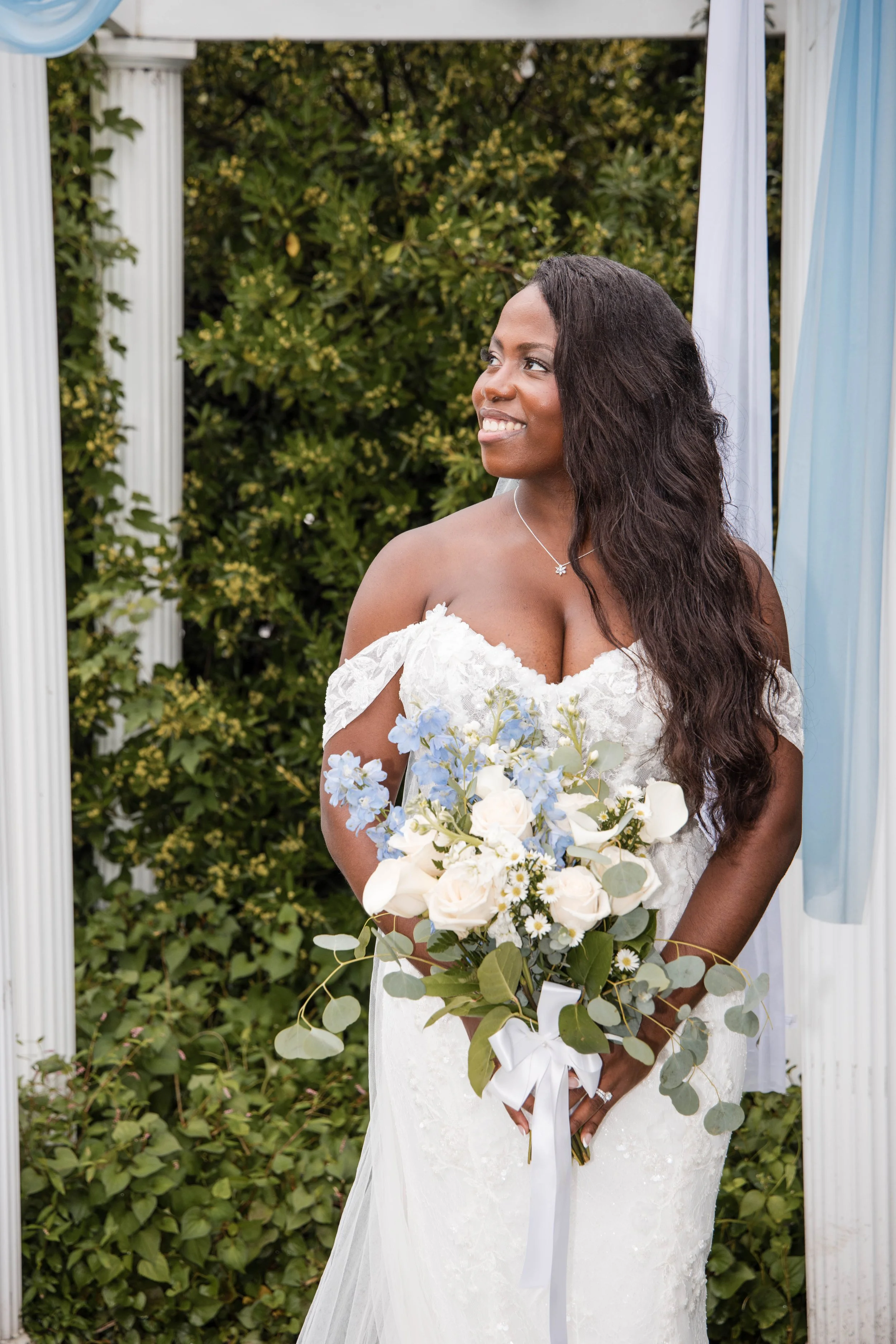 A bride with dark wavy hair, wearing a white lace wedding dress, holding a bouquet of white and light blue flowers, standing outdoors under a decorative arch with columns and light blue curtains, against a background of green foliage.