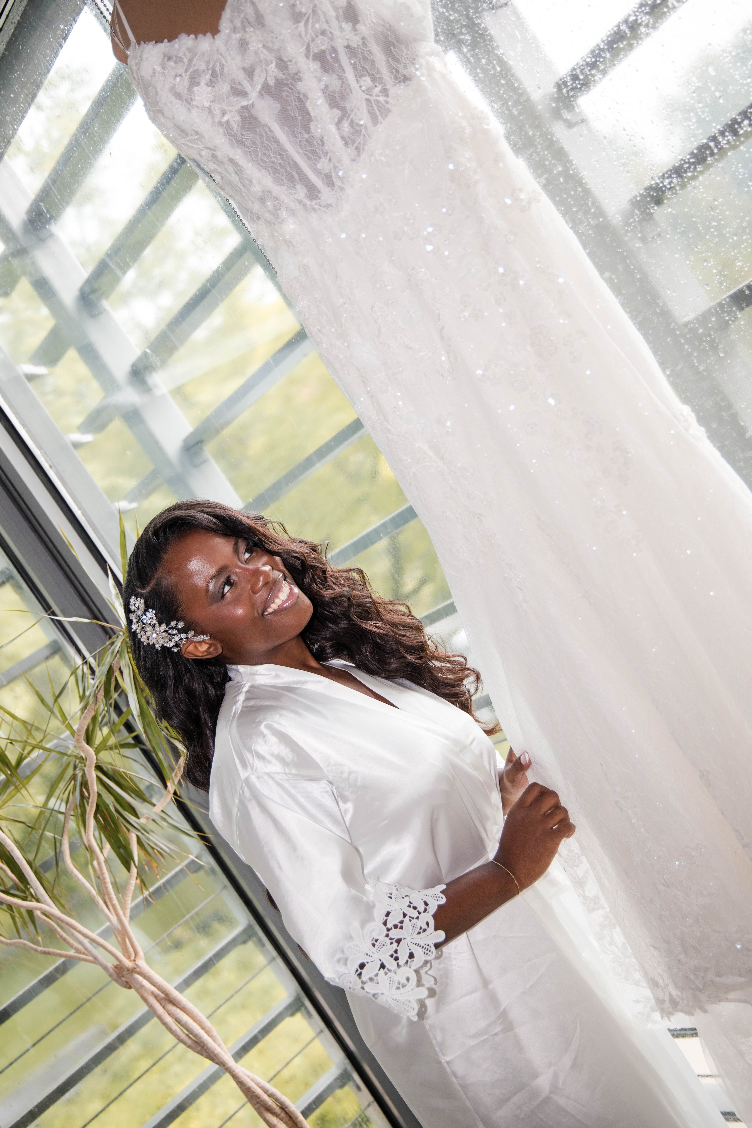 A woman with dark curly hair in a white satin robe with lace details on the sleeves, standing next to a window with a view of green trees and plants outside. She is smiling and looking to the side.