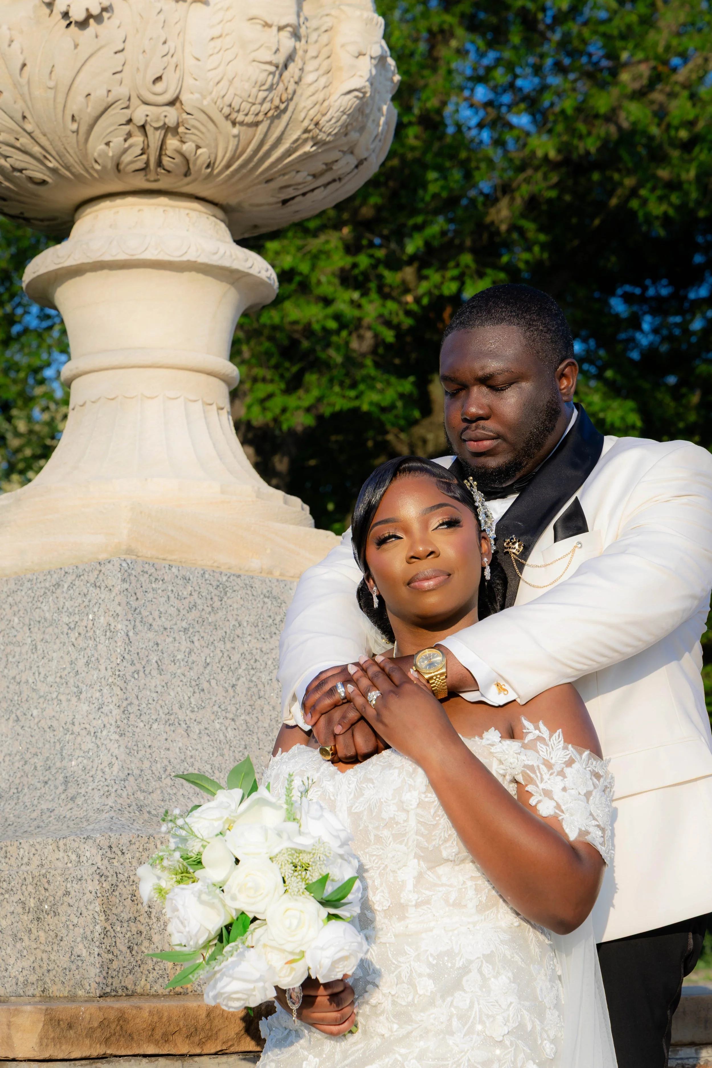 A bride and groom pose closely together outdoors, the bride holding a bouquet of white roses, with a large ornate stone flower pot and lush green trees in the background.
