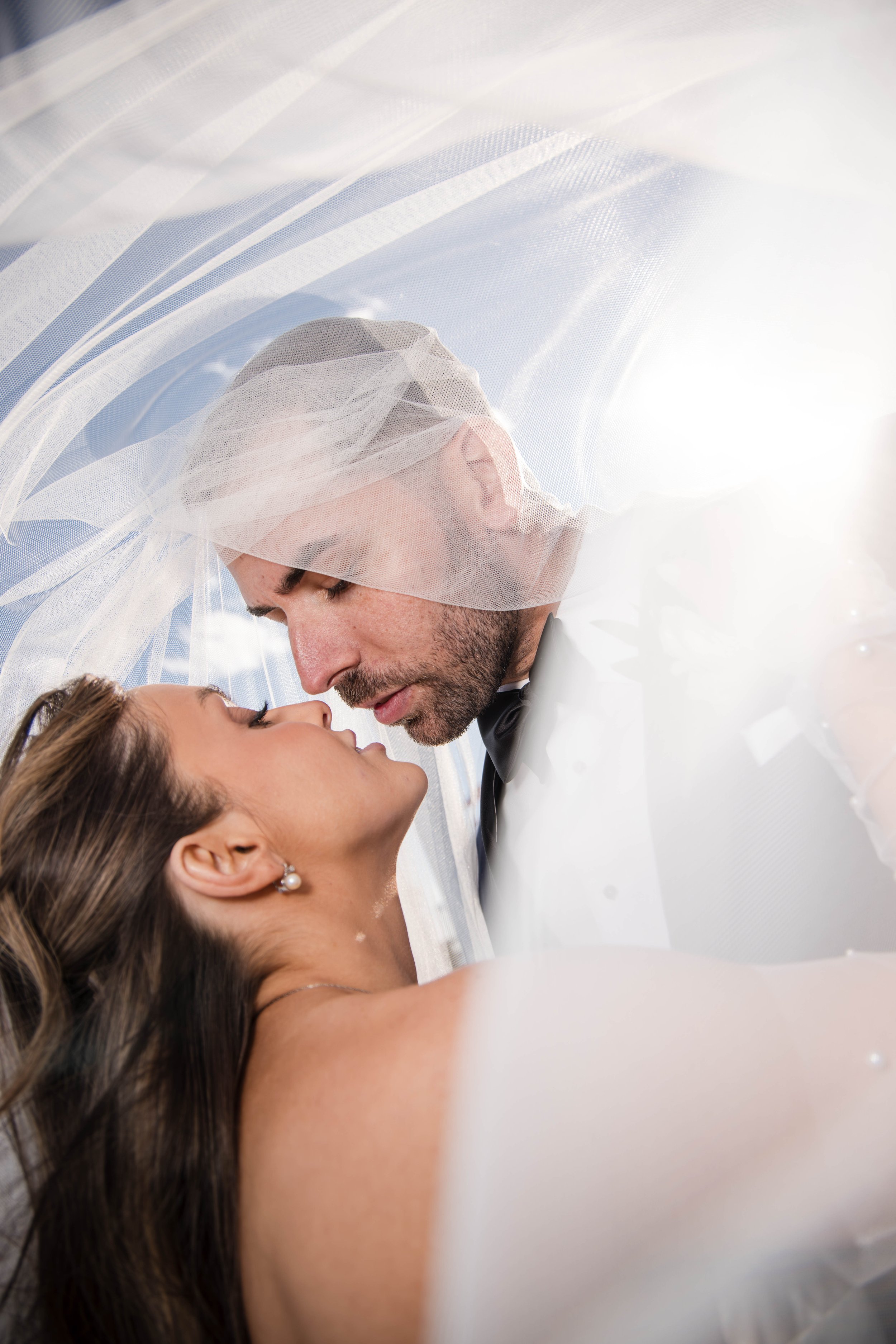 A bride and groom are about to kiss under a sheer veil at their wedding, with their faces close and eyes closed, surrounded by white decorations.