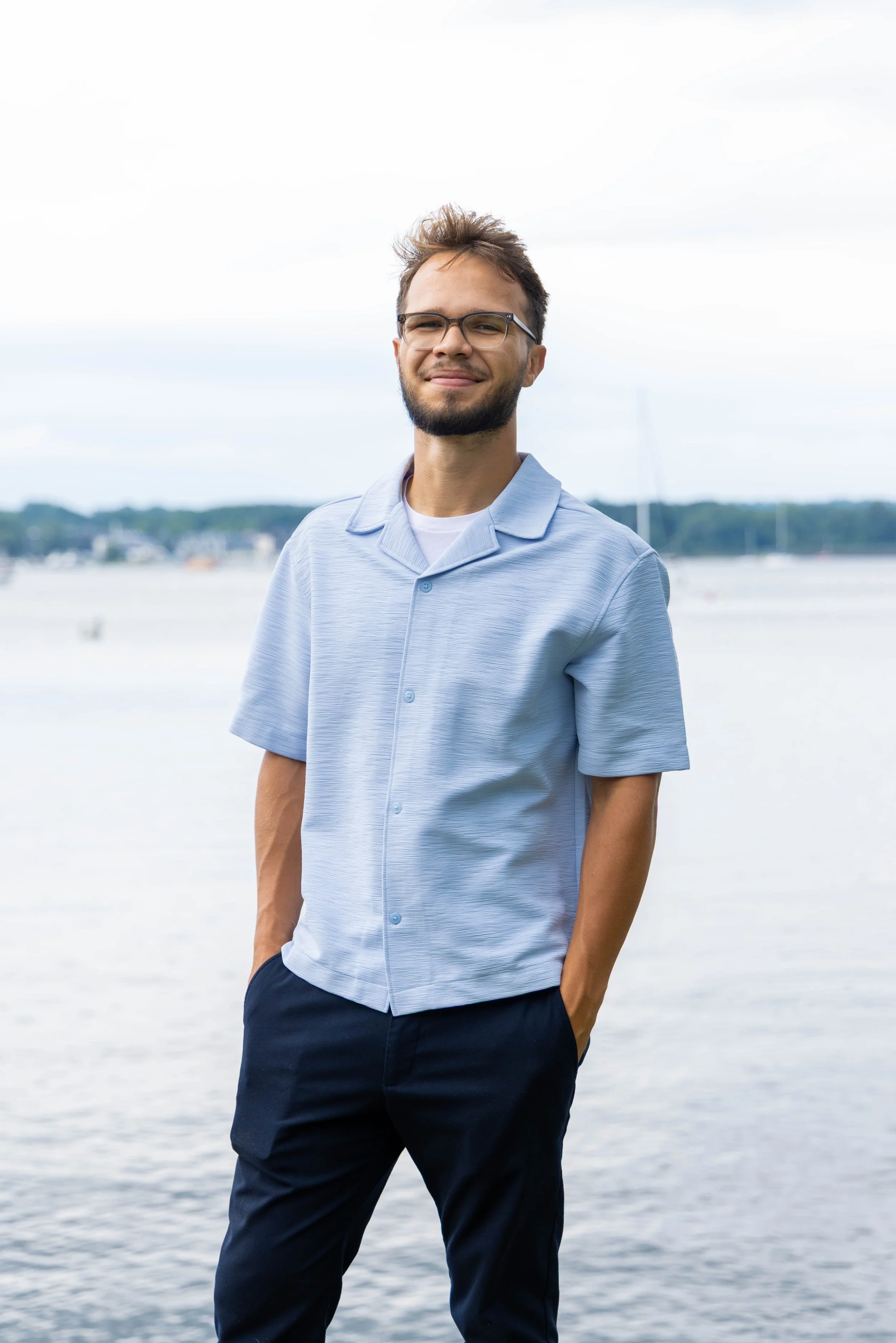 A young man with glasses, a beard, and short, messy brown hair stands near a body of water, smiling slightly. He is wearing a light blue short-sleeved button-up shirt and dark pants. The background features a cloudy sky and distant shoreline with boats.