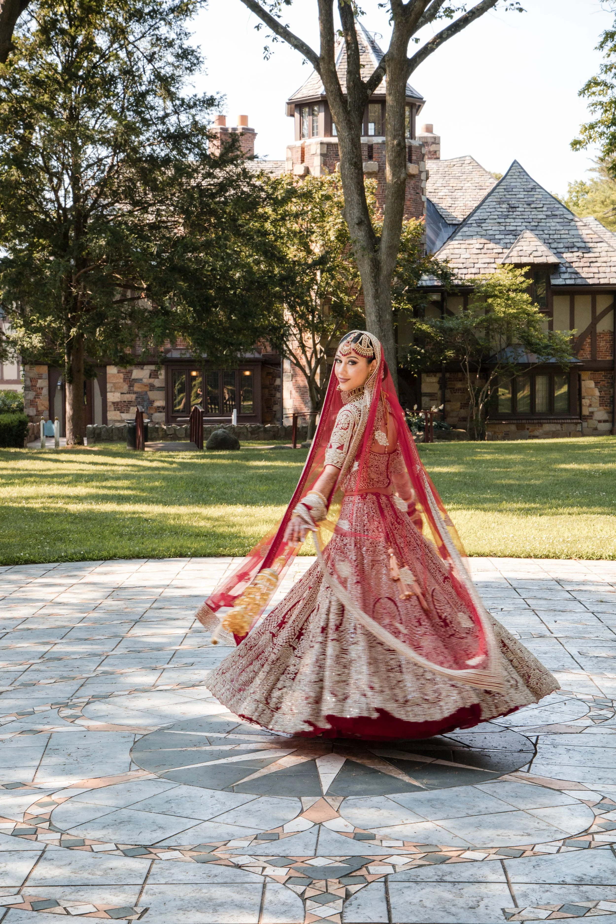 A woman dressed in traditional Indian wedding attire standing outdoors in front of a large stone house with tall trees, smiling at the camera.