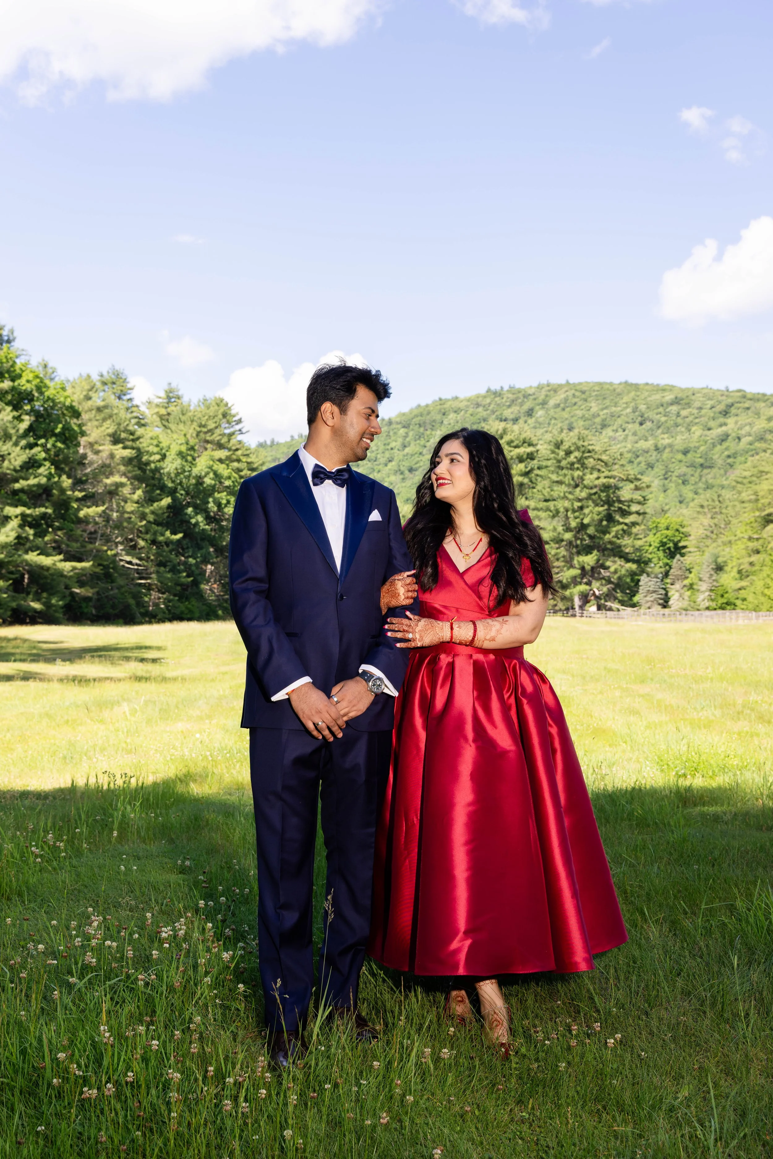 A man and a woman dressed in formal attire standing outdoors in a grassy field with trees and hills in the background, smiling at each other.