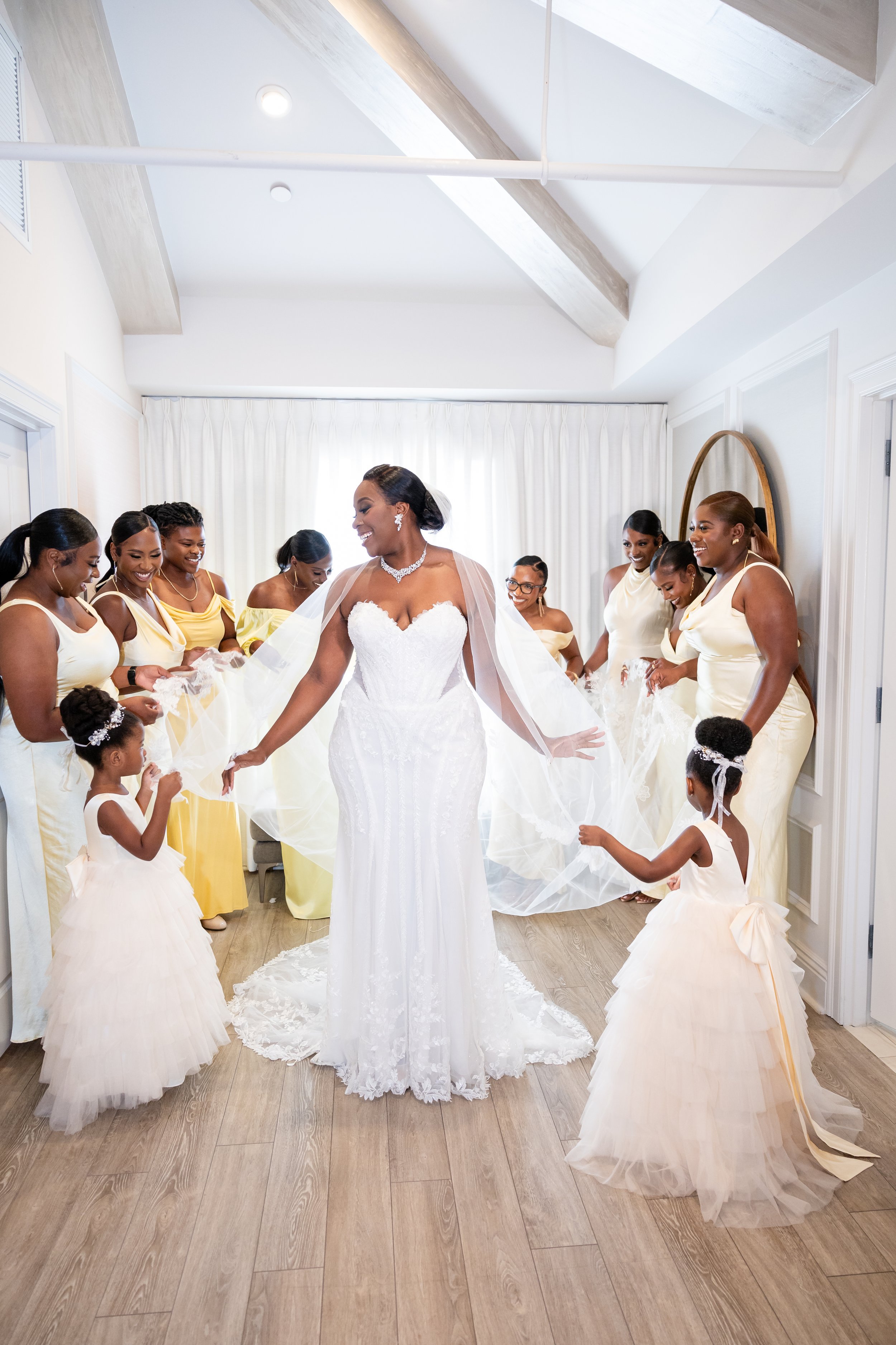A bride in a white wedding gown surrounded by bridesmaids in yellow dresses, all smiling and holding the bride's train in a bright room with wooden flooring and white curtains.