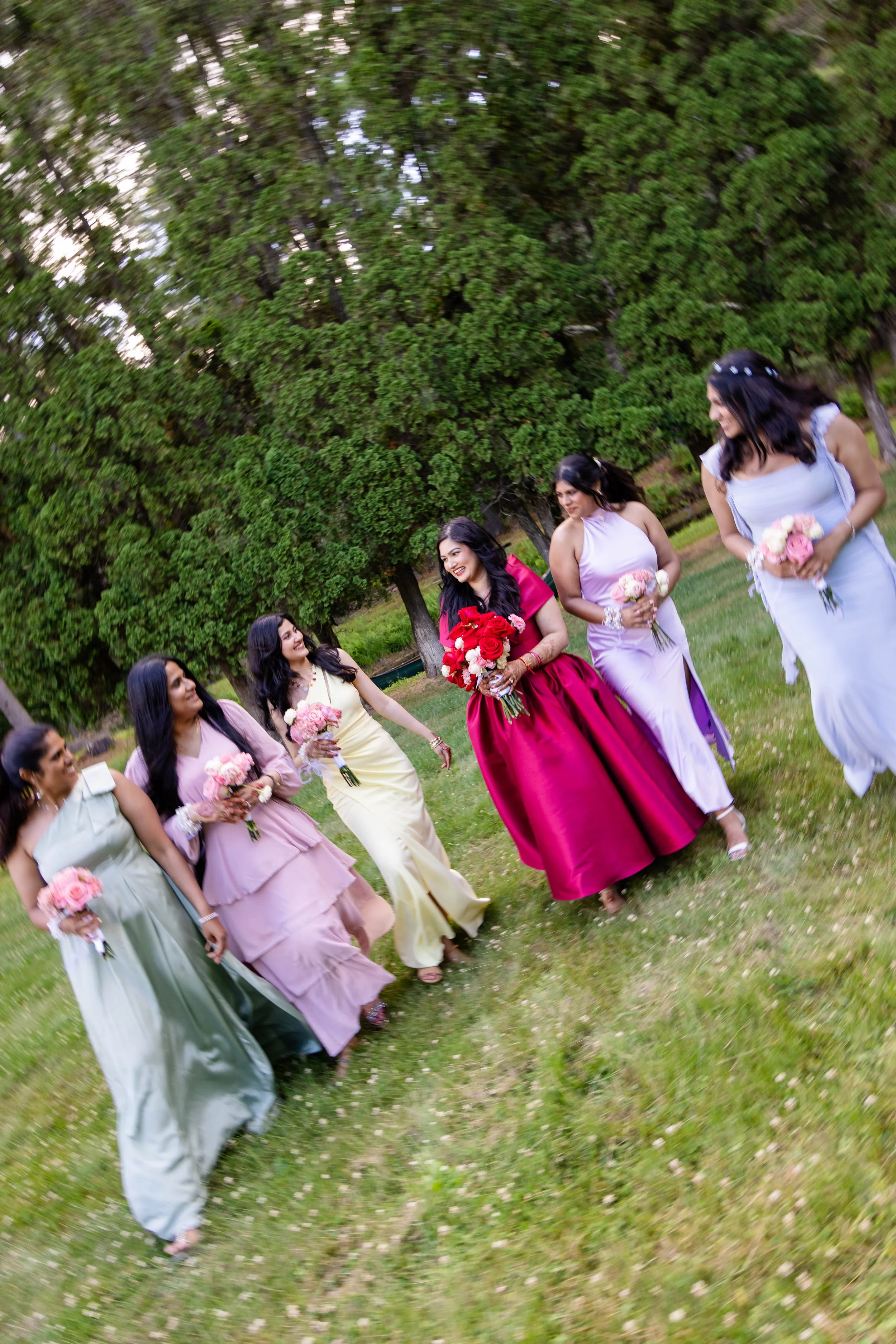 A group of women in colorful dresses holding bouquets of flowers, walking outdoors in a park or garden with green trees in the background.