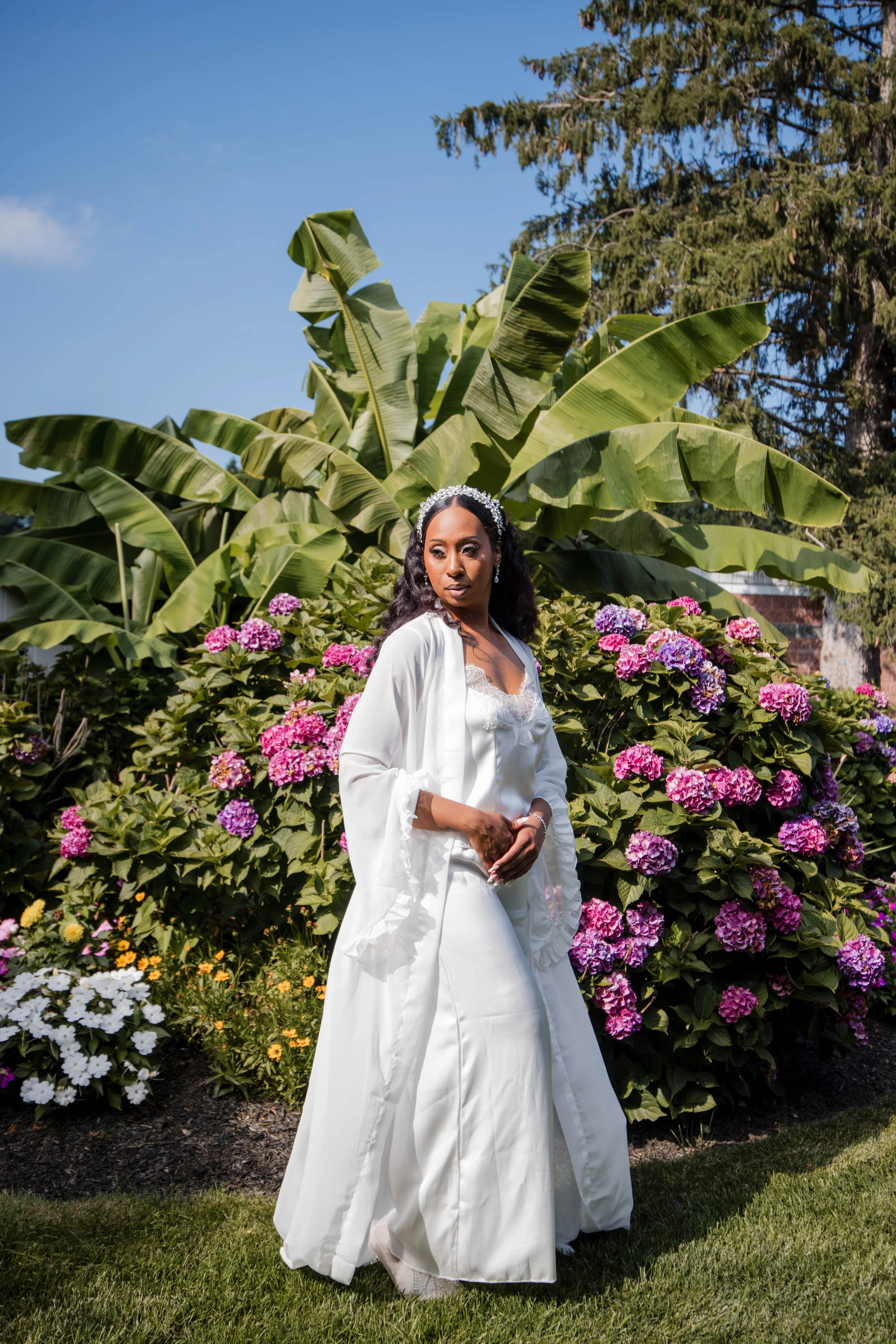 A woman dressed in wedding attire standing in a garden with flowering bushes and large green leafy plants. She is wearing a white gown with a satin finish, a lace bodice, and a white satin wrap or cape. She has dark curly hair, jewelry, and a floral headpiece, and she is looking off to the side with a serious expression.