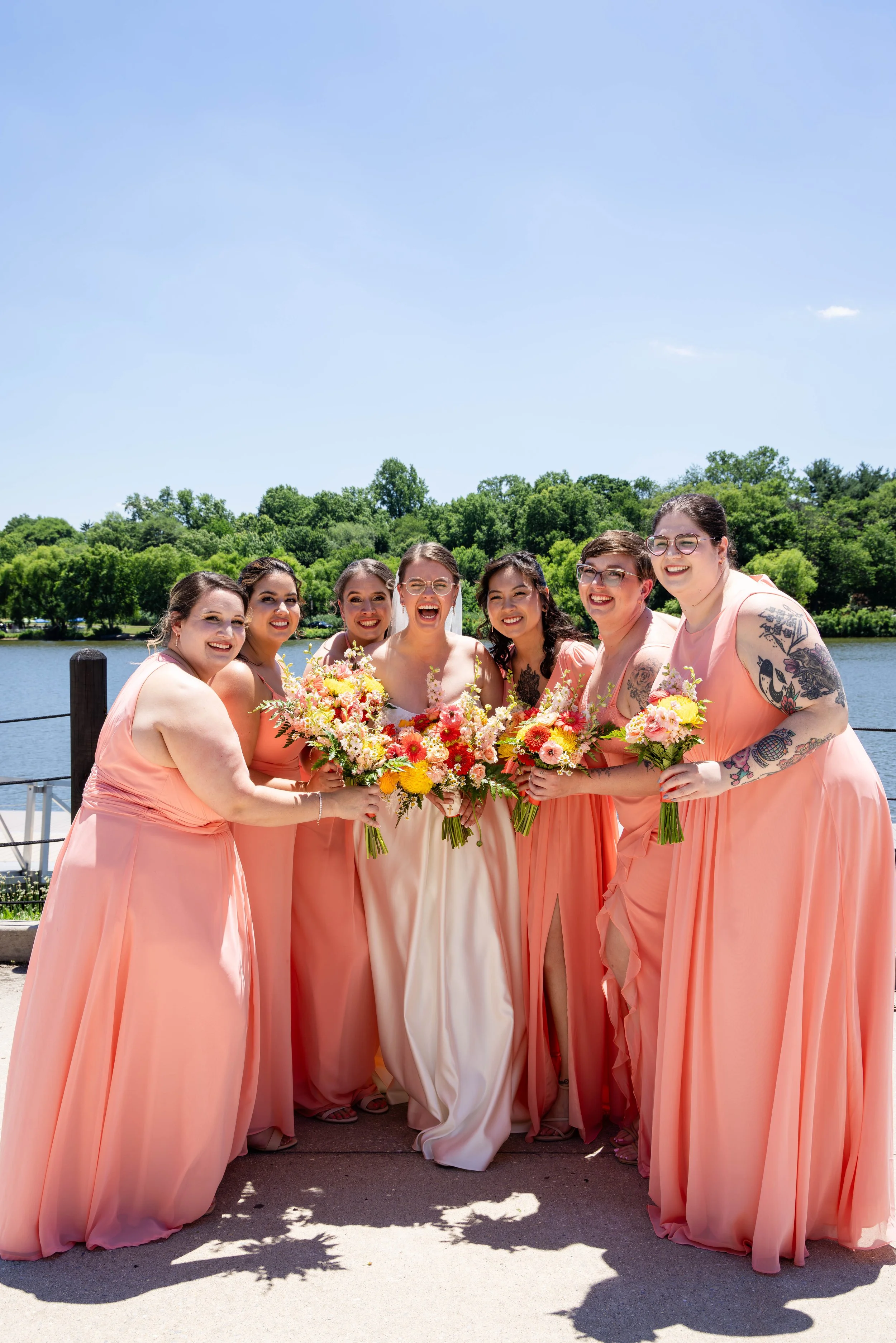 A group of eight women in pink dresses standing outdoors near a body of water, holding bouquets of flowers, smiling, with trees and a blue sky in the background.