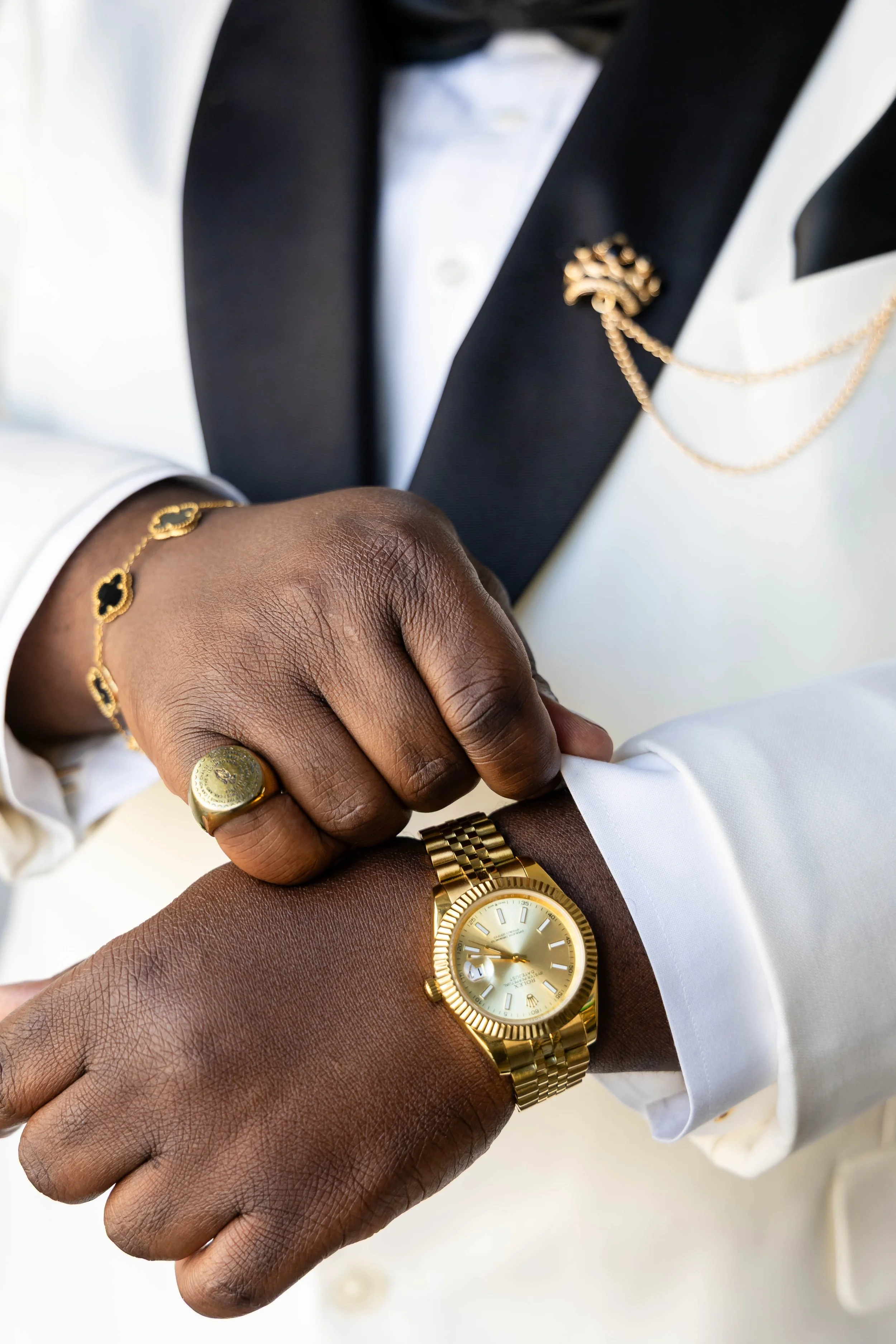 Close-up of a person with dark skin wearing a gold watch, gold bracelet, and gold ring, adjusting the sleeve of a white shirt. Another person with darker skin is also wearing a gold bracelet, and a suit jacket with a gold chain pin is visible.