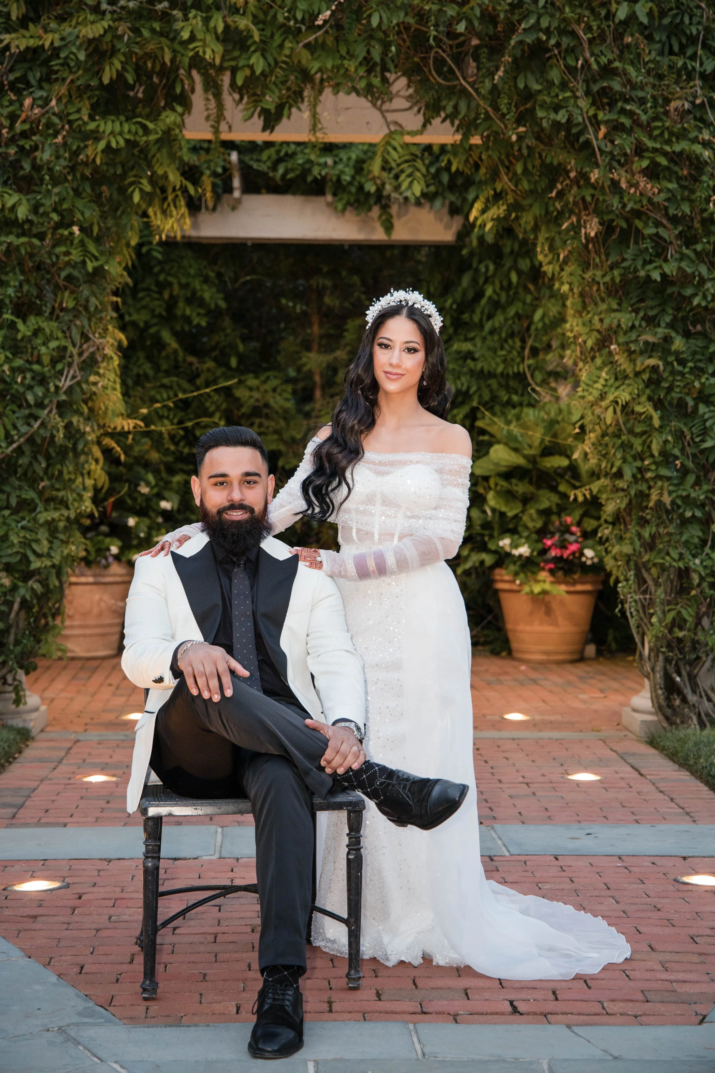 A bride and groom pose together outdoors, surrounded by greenery. The groom is seated on a chair, dressed in a white tuxedo with black lapels, black shirt, and black tie. The bride stands beside him, wearing a white off-the-shoulder wedding gown with lace details and a tiara. They are smiling, and the setting features potted plants and a trellis arch.