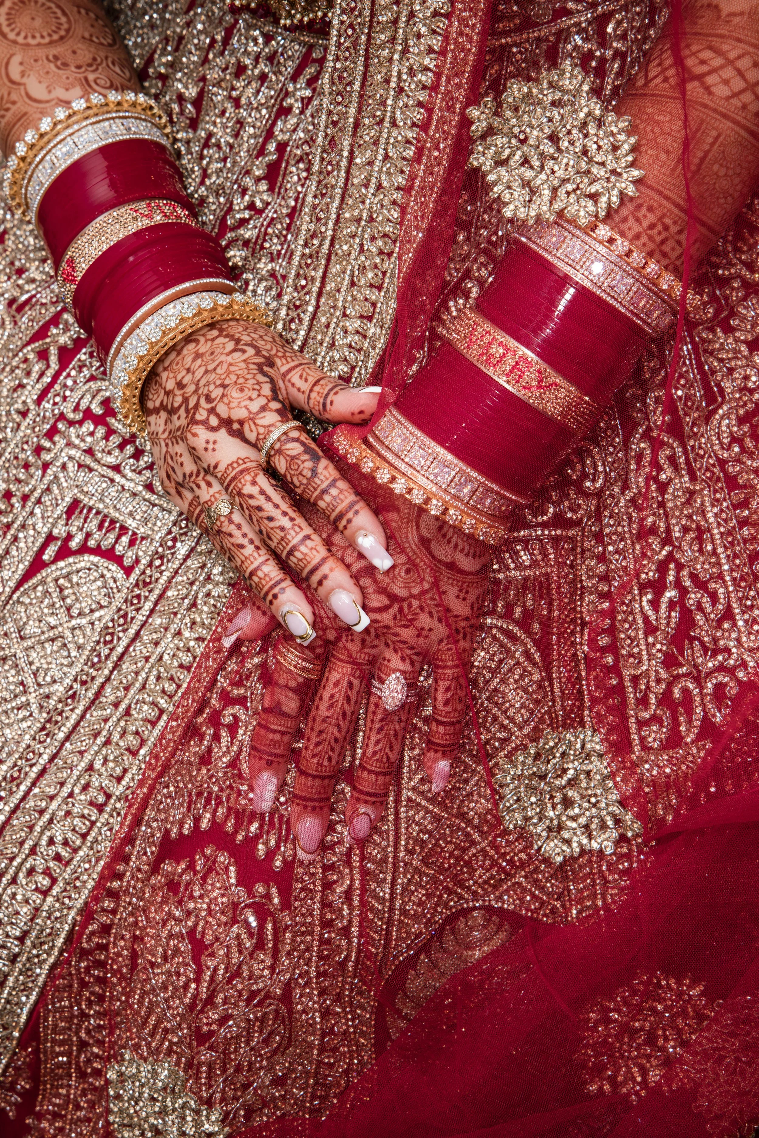 Close-up of a woman wearing a red and gold heavily embroidered traditional Indian saree with intricate patterns and embellishments, gold jewelry, bangles, and henna tattoos on her hand.