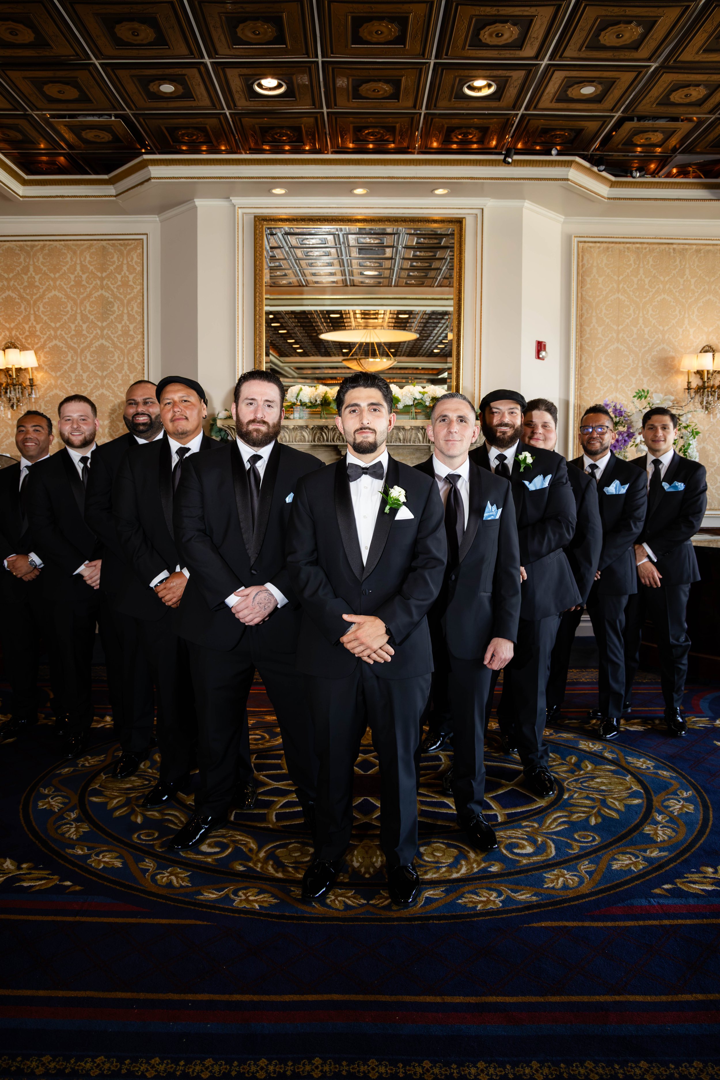 Group of well-dressed men in tuxedos standing in a formal room with ornate ceiling, mirror, floral arrangements, and patterned carpet.