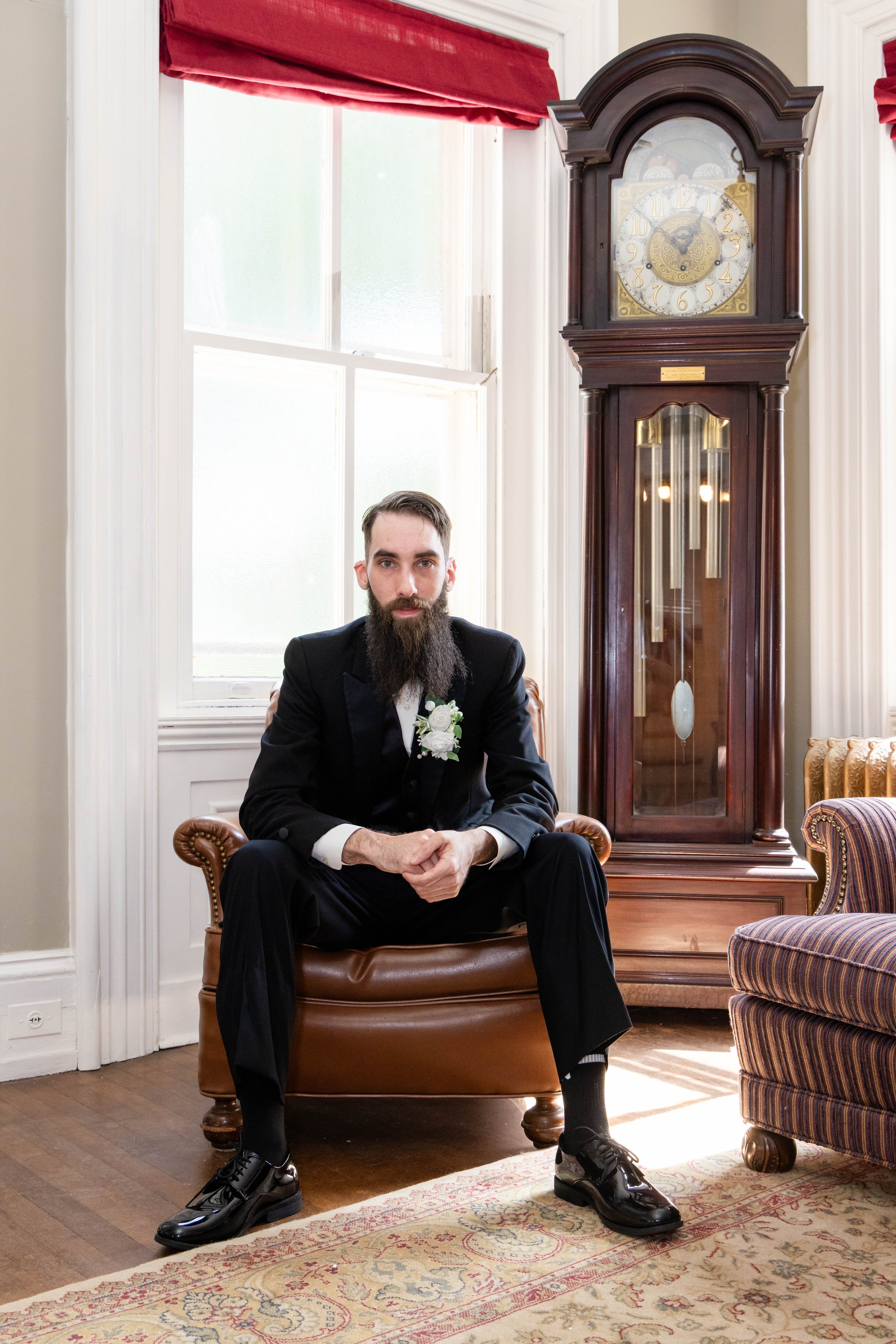 A man in a tuxedo with a beard sitting on a brown leather armchair in a room with large windows and red curtains, next to a tall wooden grandfather clock and an upholstered striped armchair.