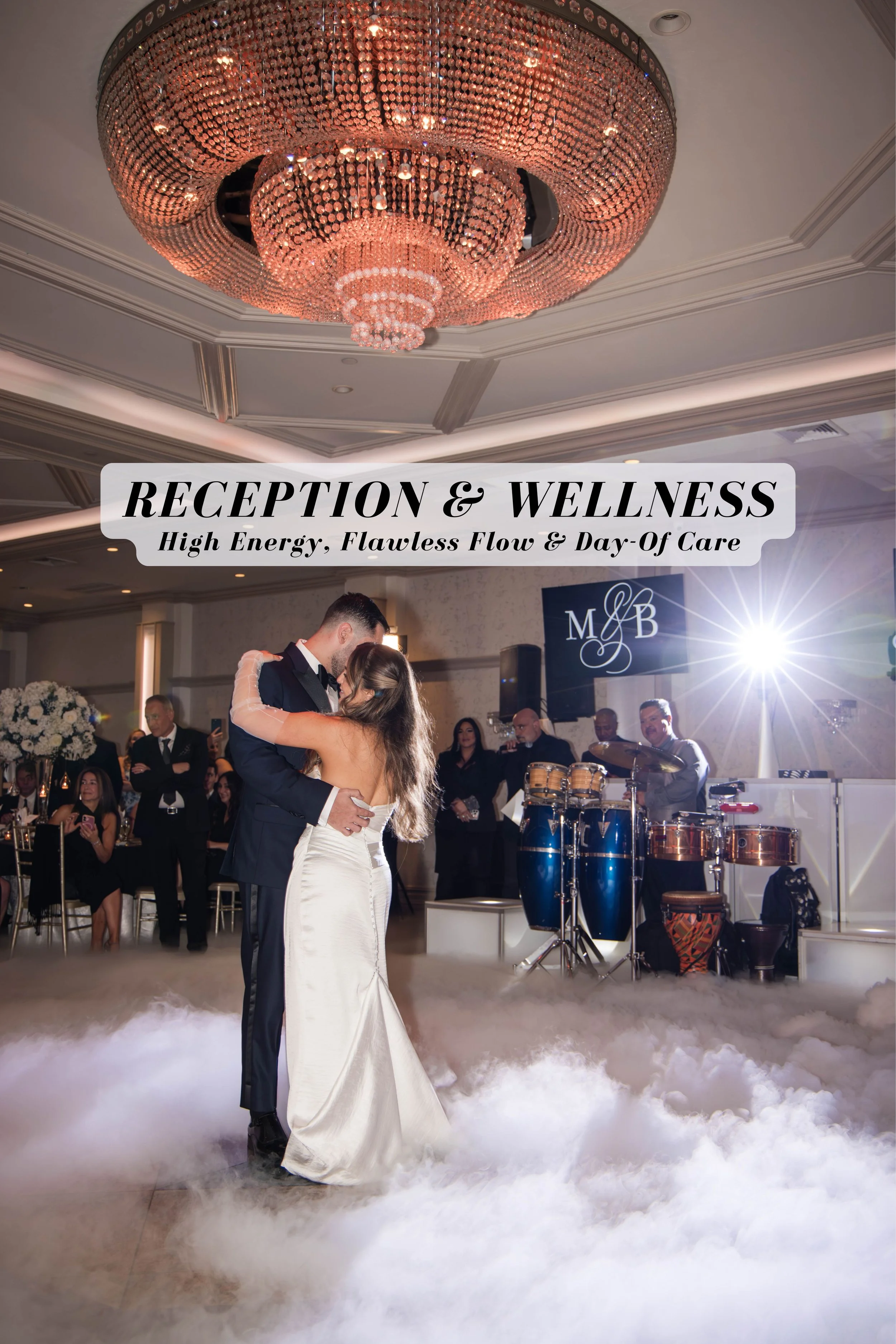A bride and groom dancing at their wedding reception, surrounded by guests and a live band with drums. The ceiling features a large, elegant chandelier, and the dance floor is covered with a cloud-like fog.