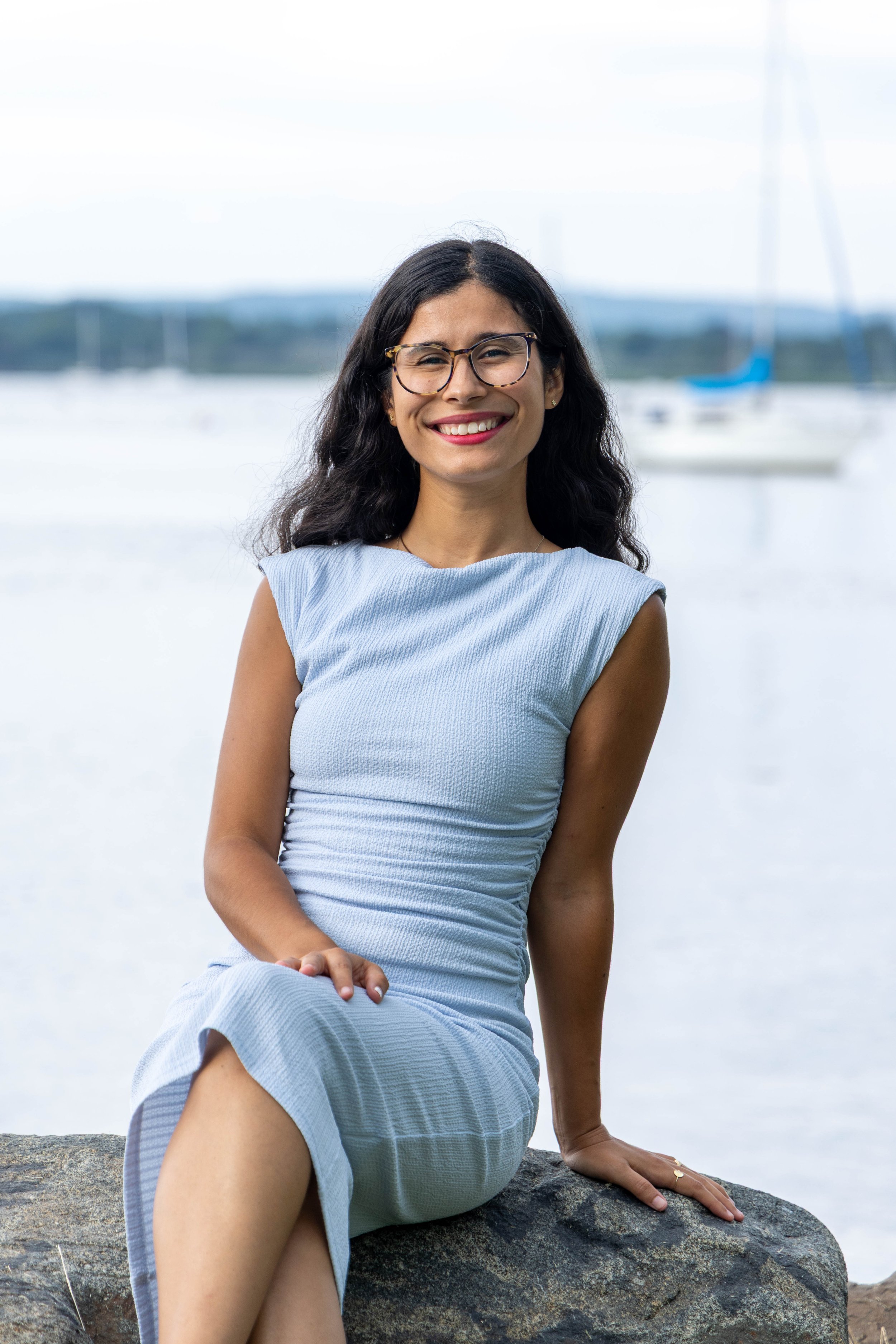 A woman with long dark hair and glasses smiling while sitting on a rock near the water with sailboats in the background.
