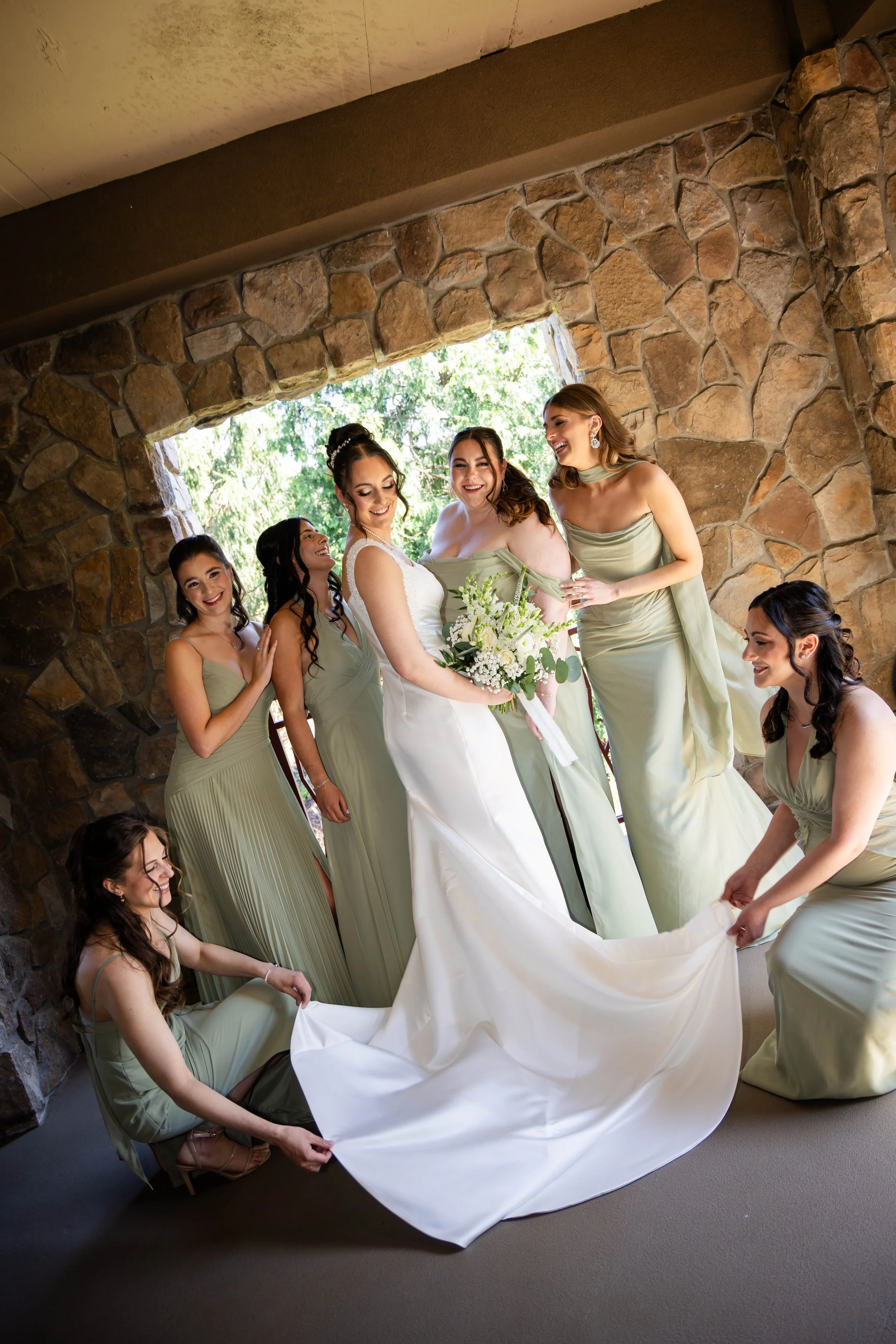 Bride in a white wedding dress holding a bouquet of white flowers and greenery, surrounded by smiling bridesmaids in matching light green dresses with pleated skirts at a stone wall outdoor venue.