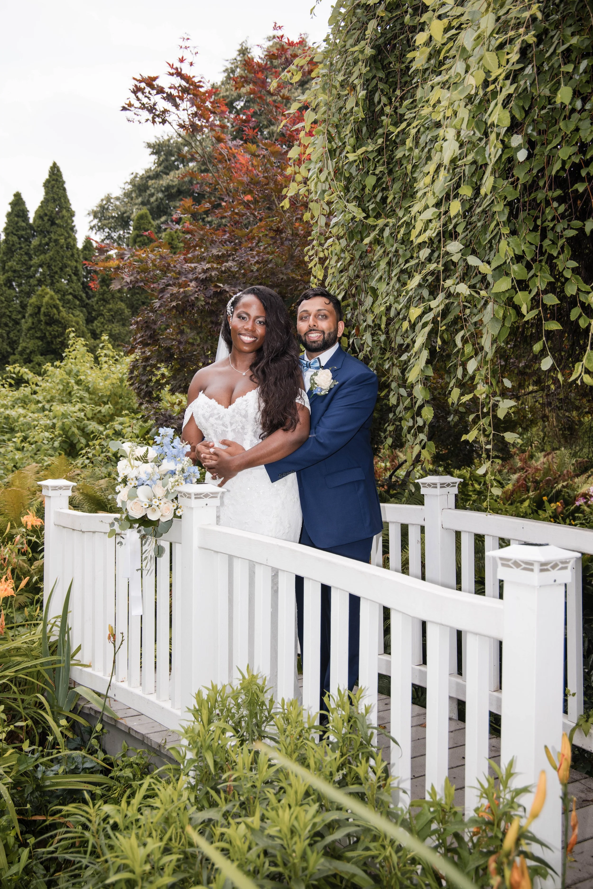 A bride and groom standing close together outdoors in front of a white picket fence and surrounded by lush green and colorful autumn trees.