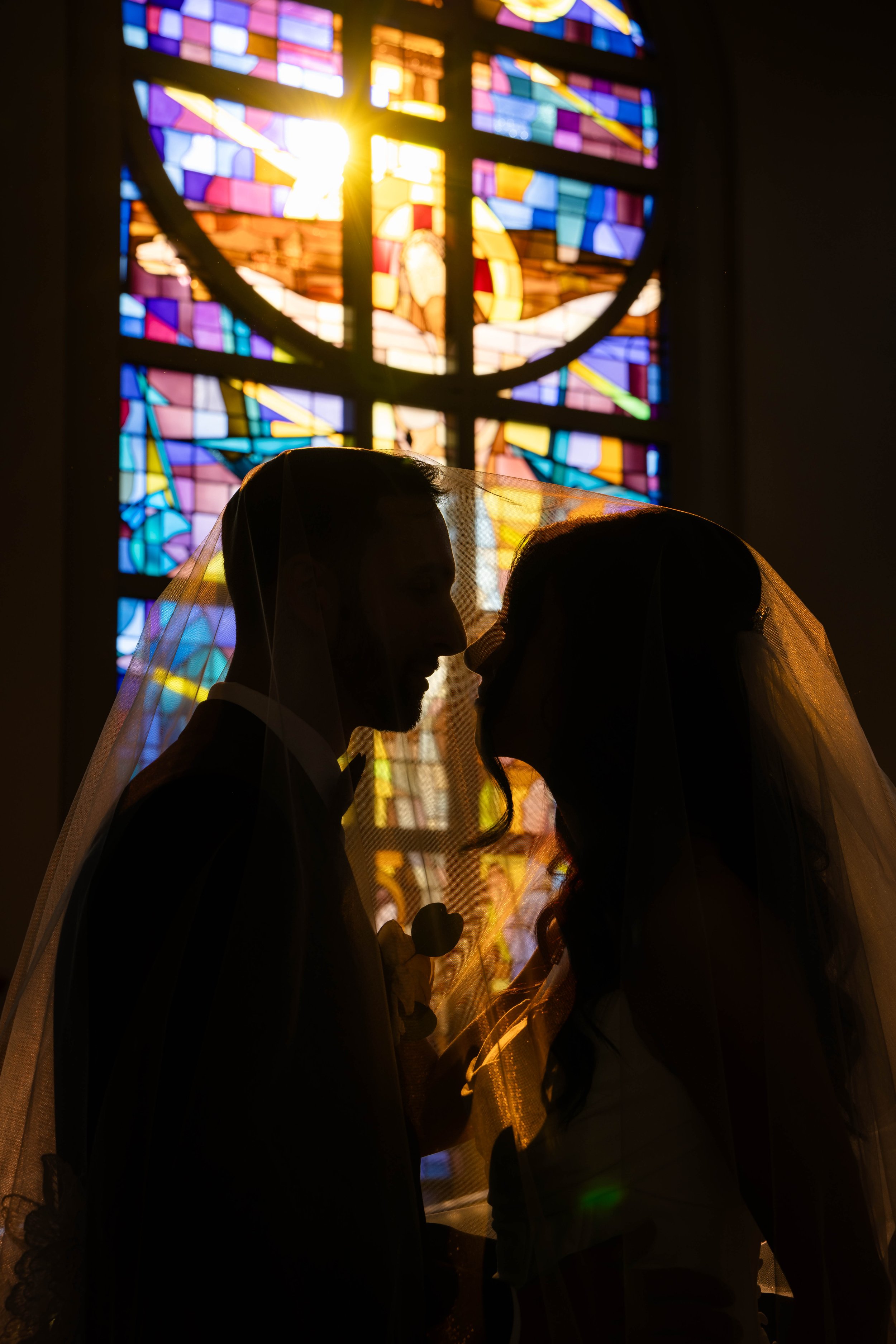 Silhouettes of a bride and groom standing close together with a stained glass window in the background, sunlight shining through.