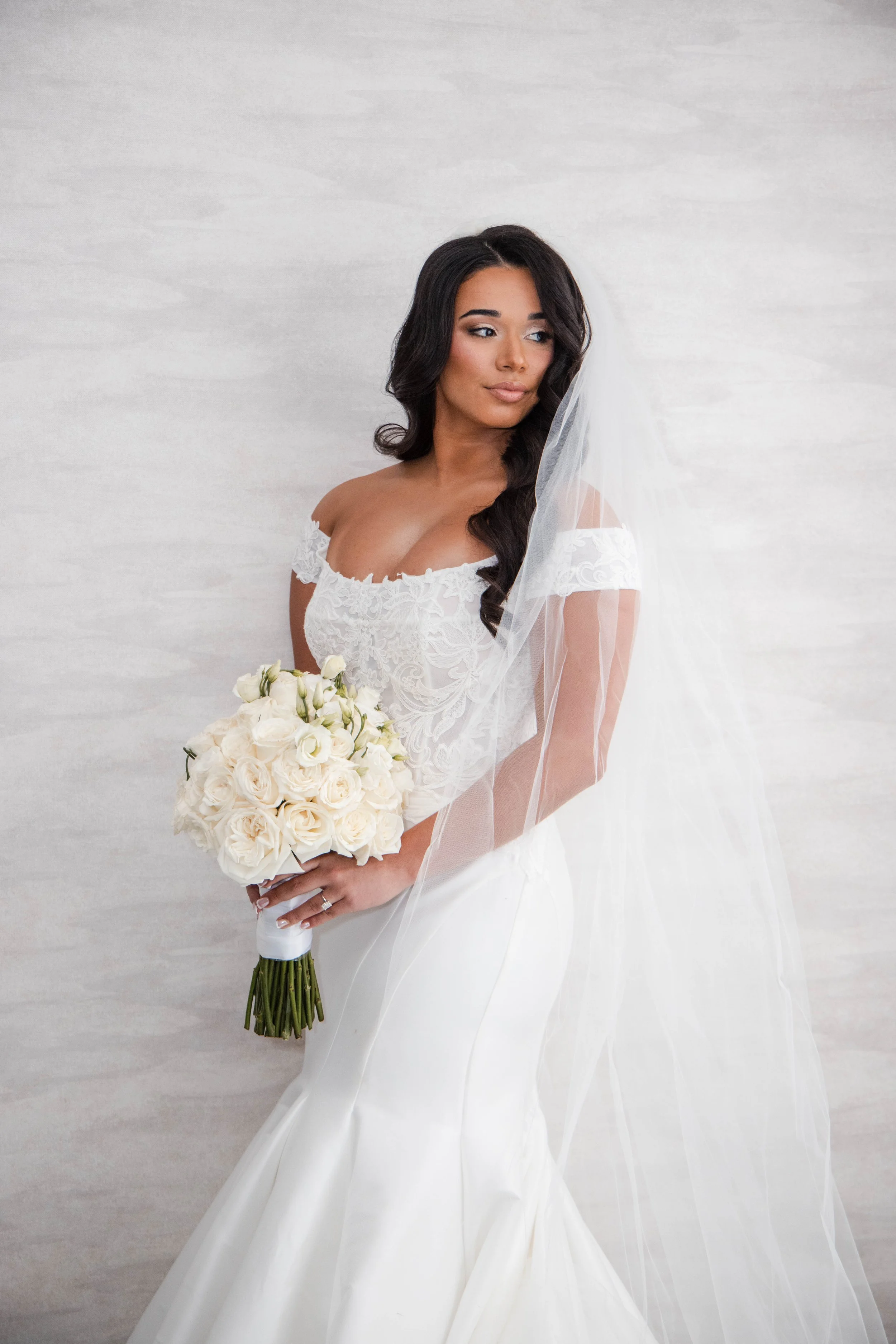 A bride in a white wedding dress with lace details, holding a bouquet of white roses, and wearing a veil, standing against a light-colored textured background.