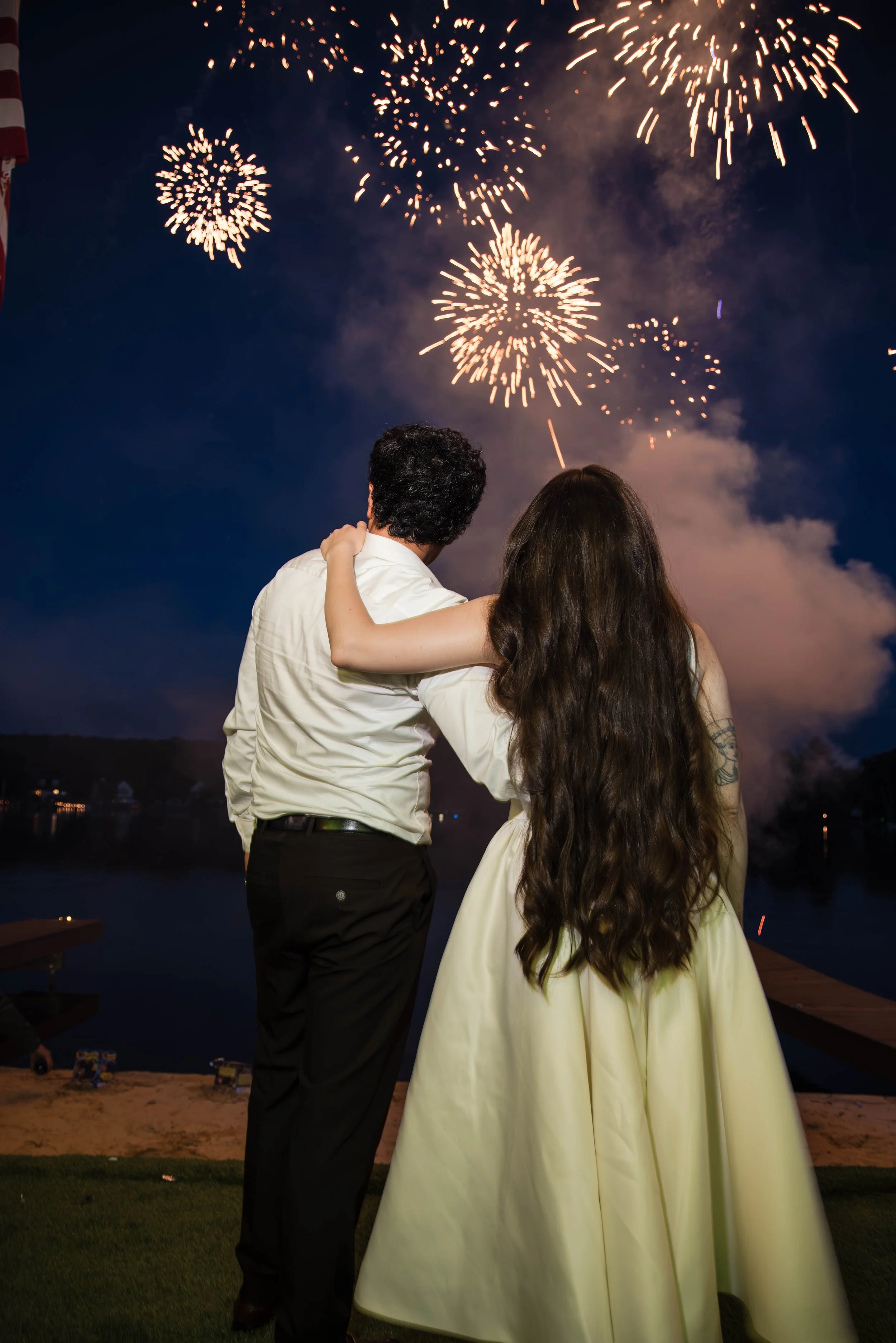 A couple watching fireworks at night, with their arms around each other. The man is wearing a white shirt and black pants, and the woman is wearing a light-colored dress. The fireworks light up the dark sky above a body of water, creating reflections, with some buildings visible in the distance.
