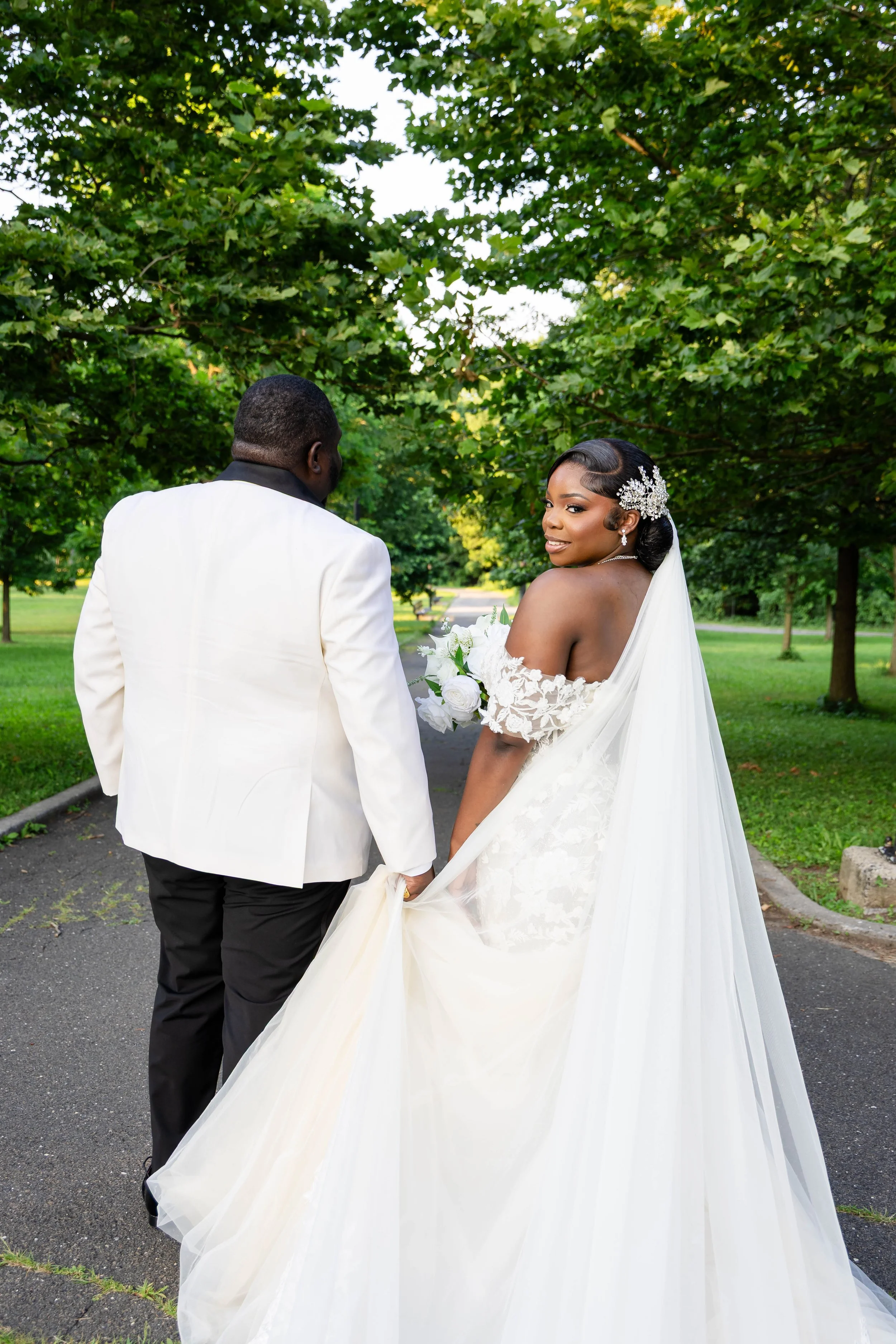 A bride and groom holding hands outdoors, with the bride smiling and wearing a white wedding gown and veil, and the groom in a white tuxedo jacket. Behind them are green trees and a pathway.