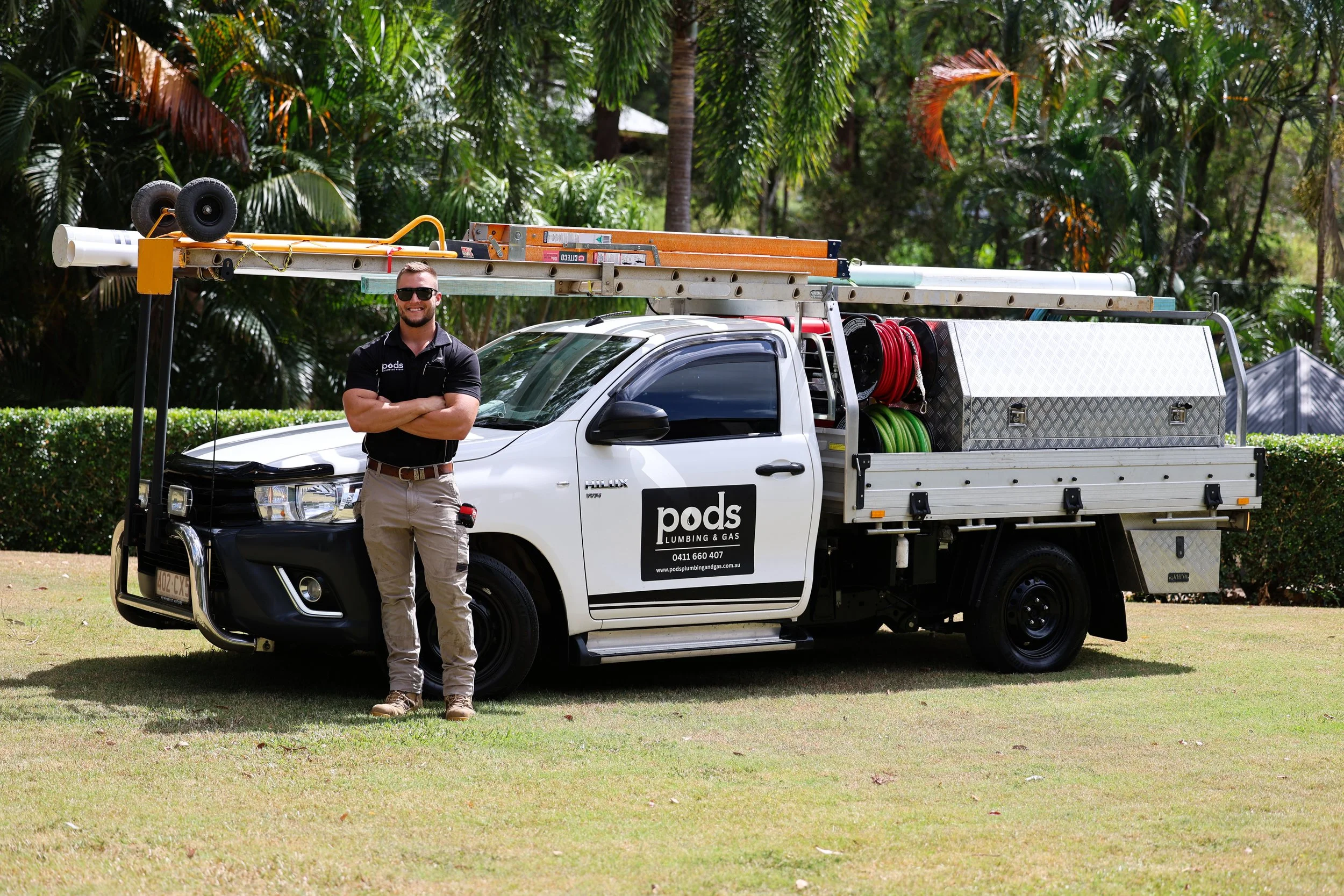 A man standing with arms crossed next to a white service truck labeled 'pods Plumbing & Gas,' parked on a grassy area with trees in the background. The truck is equipped with ladders, hoses, and storage compartments.