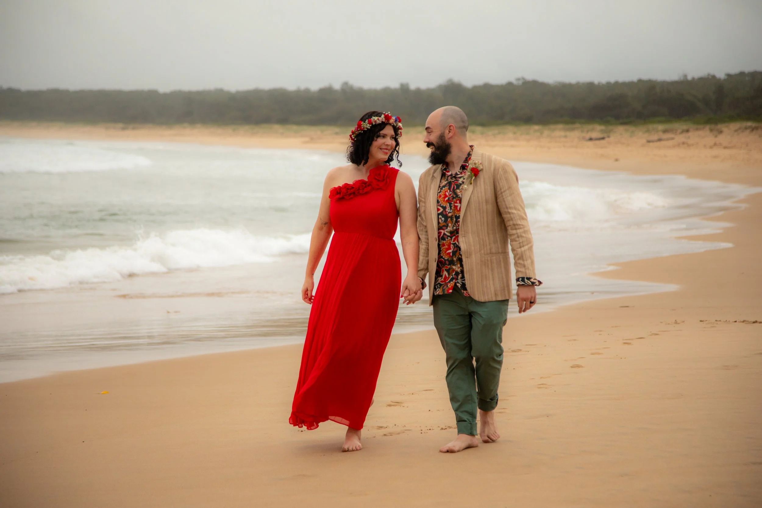 A couple walking hand-in-hand along the beach, dressed in colorful, stylish clothes, with the ocean and cloudy sky in the background.