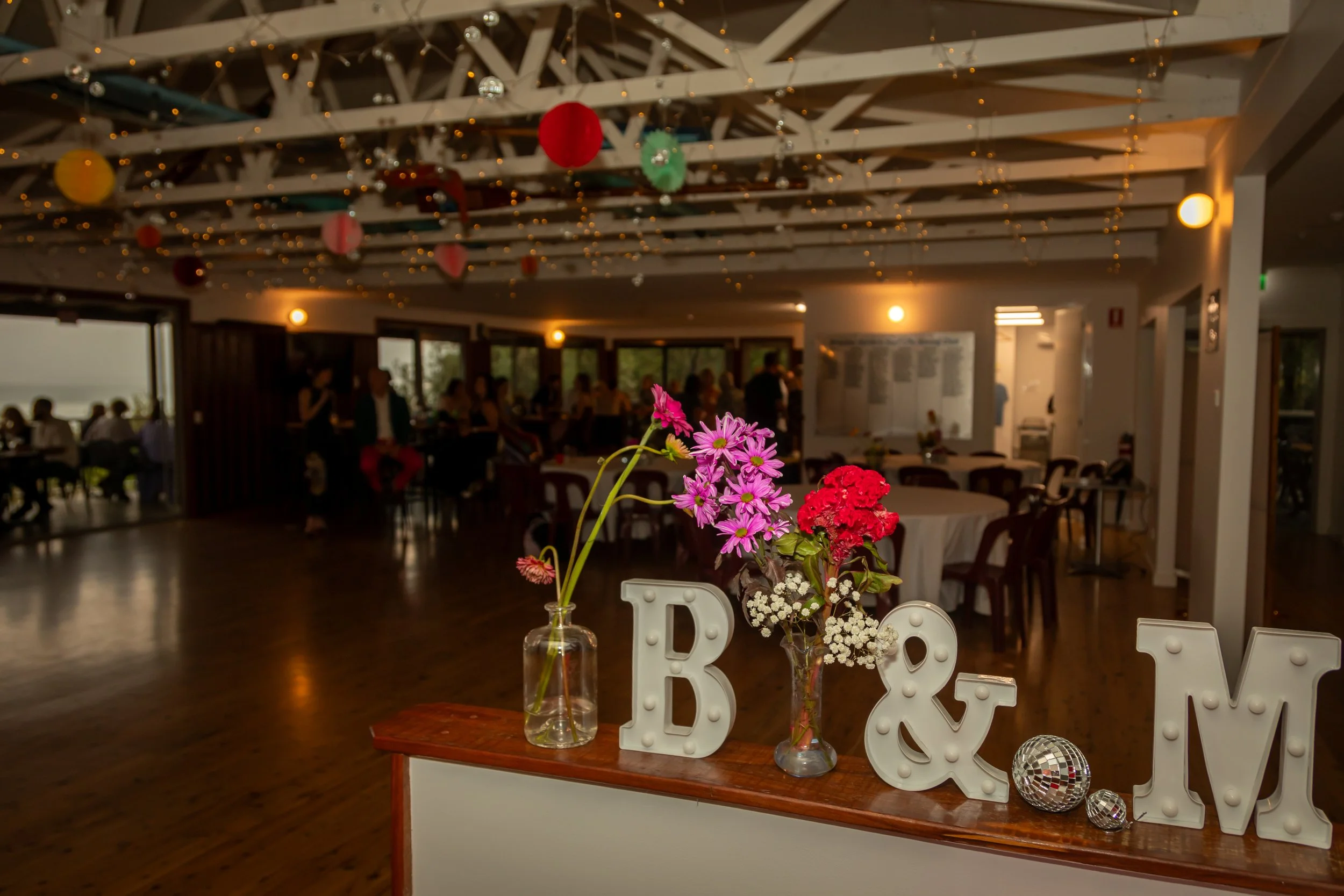 Decorated event space with pink and red flowers in vases, large white letters spelling 'B & M', and small disco balls on a wooden table. The background shows tables, chairs, and people, with colorful lanterns and string lights hanging from the ceiling.