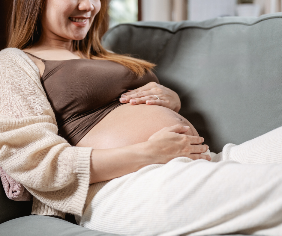 Pregnant woman sitting on a sofa, smiling and resting her hands on her belly, indoors.