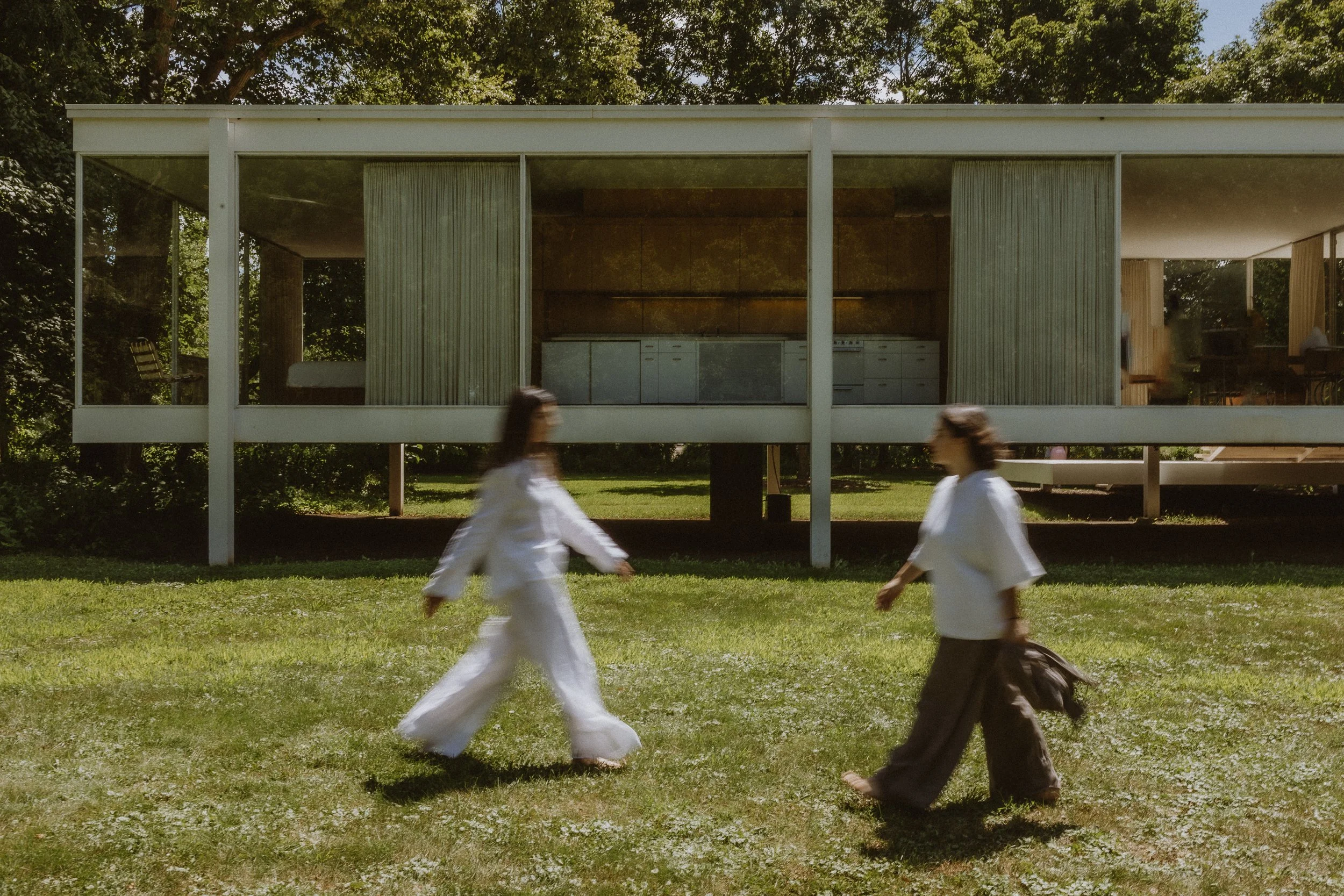 Two women walking across a grassy area in front of a modern, glass-fronted house with trees in the background.