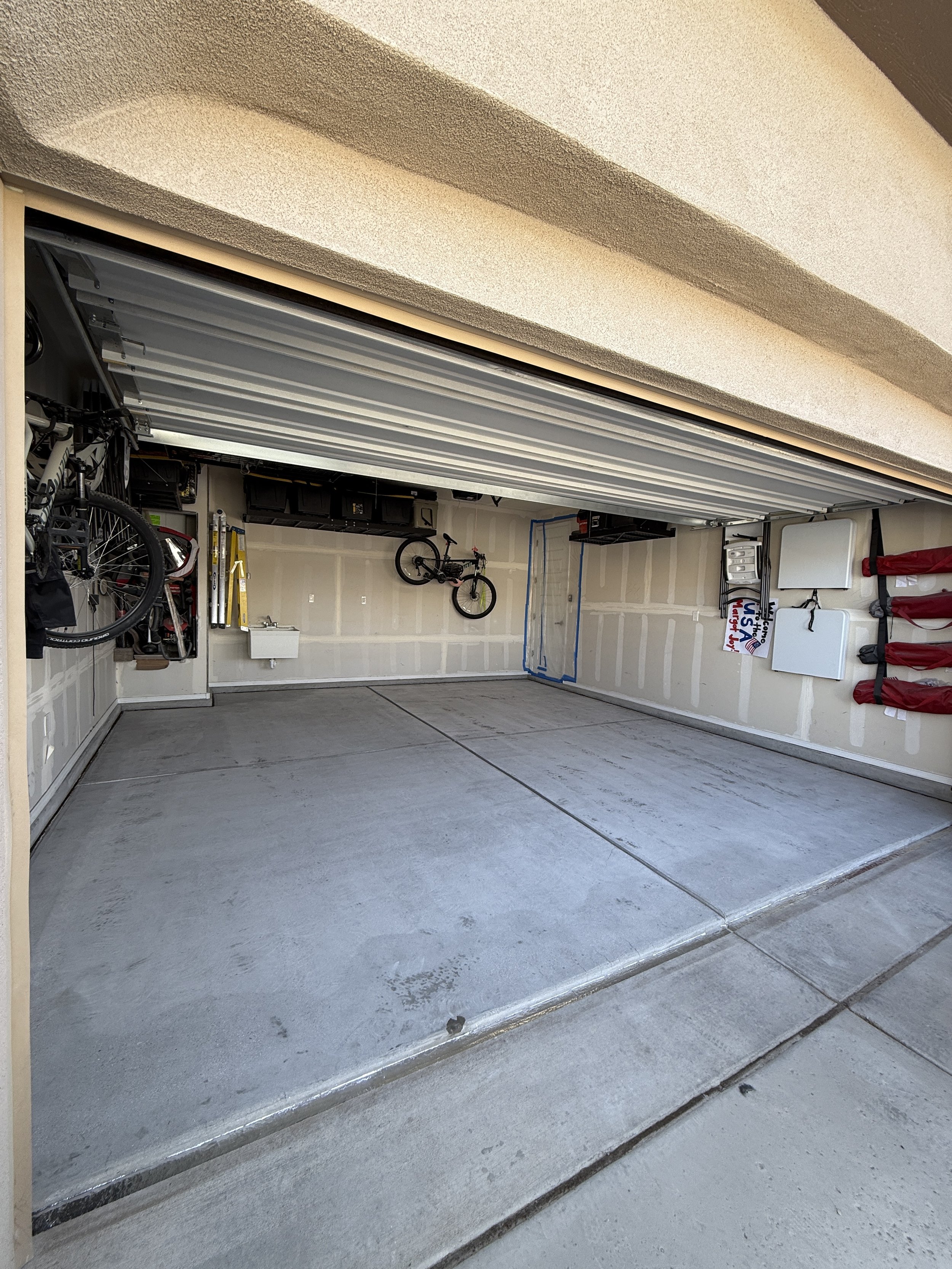 Open garage with concrete floor, bicycle hanging on the wall, storage shelves above, and various items organized along the sides.