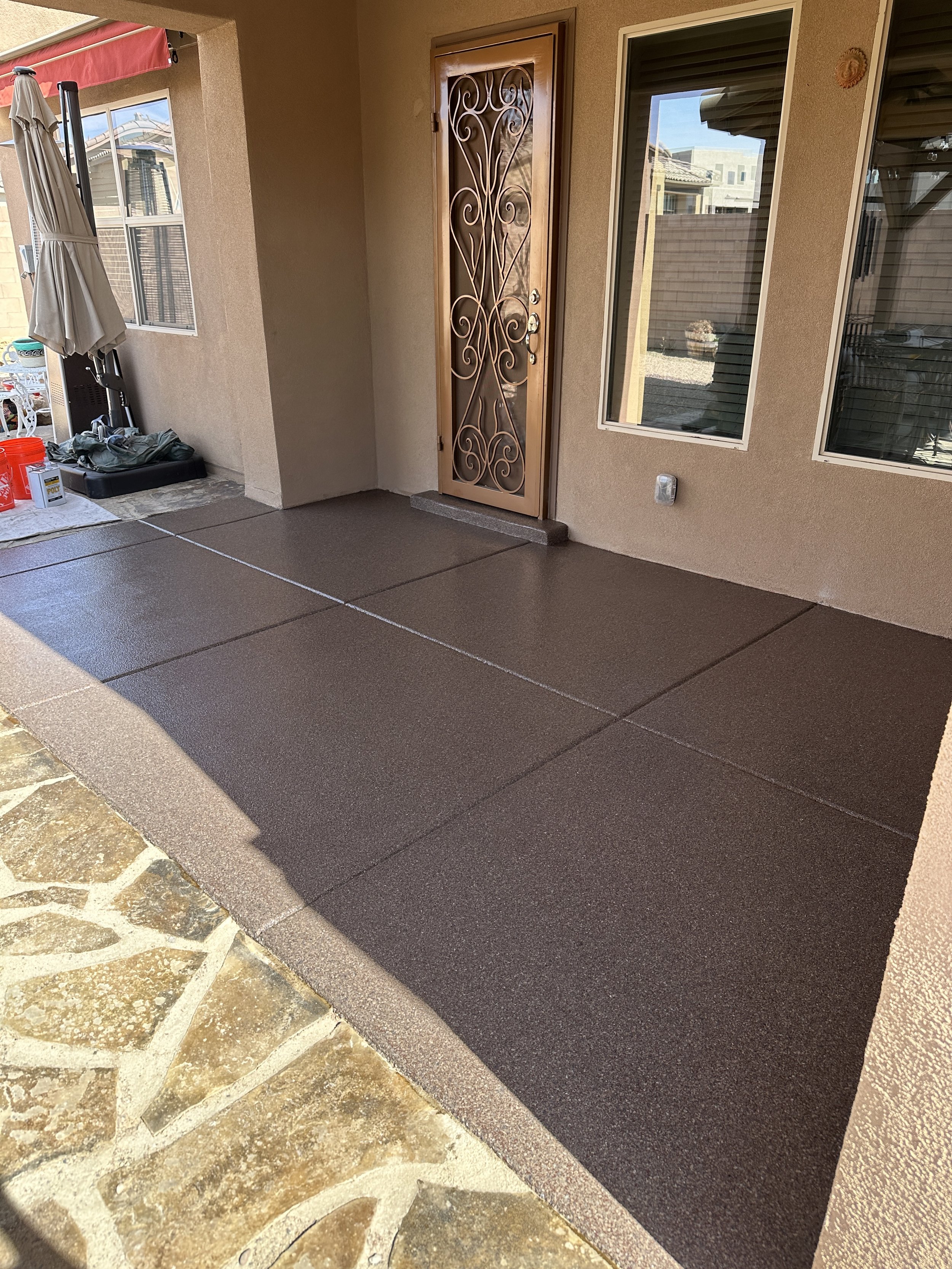 A residential patio with a decorative front door, two large windows, an umbrella, and neatly tiled flooring.