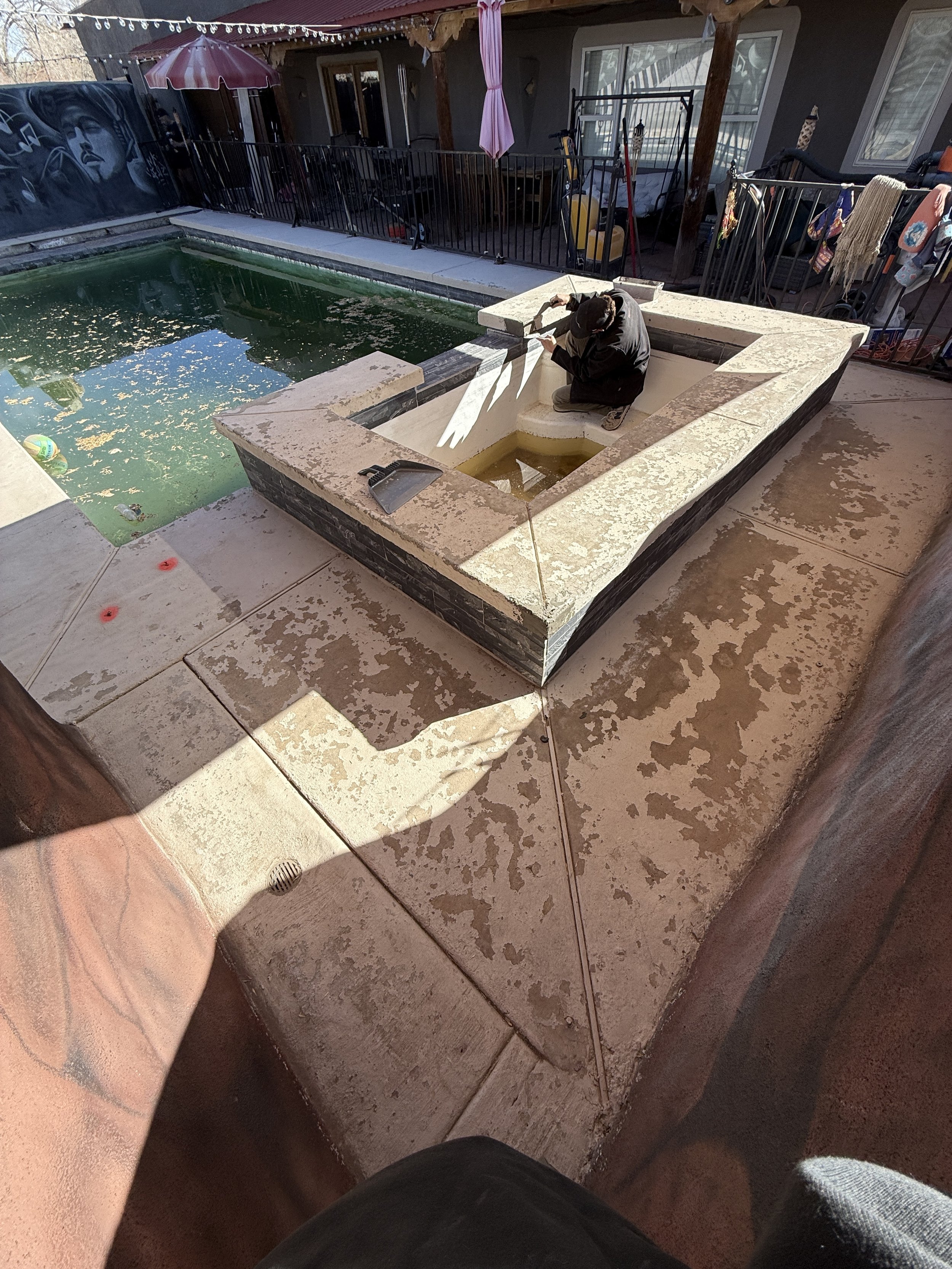 Outdoor pool area with a small empty hot tub being repaired by a person, surrounded by a fence, with fallen leaves in the pool water and a mural on a wall nearby.