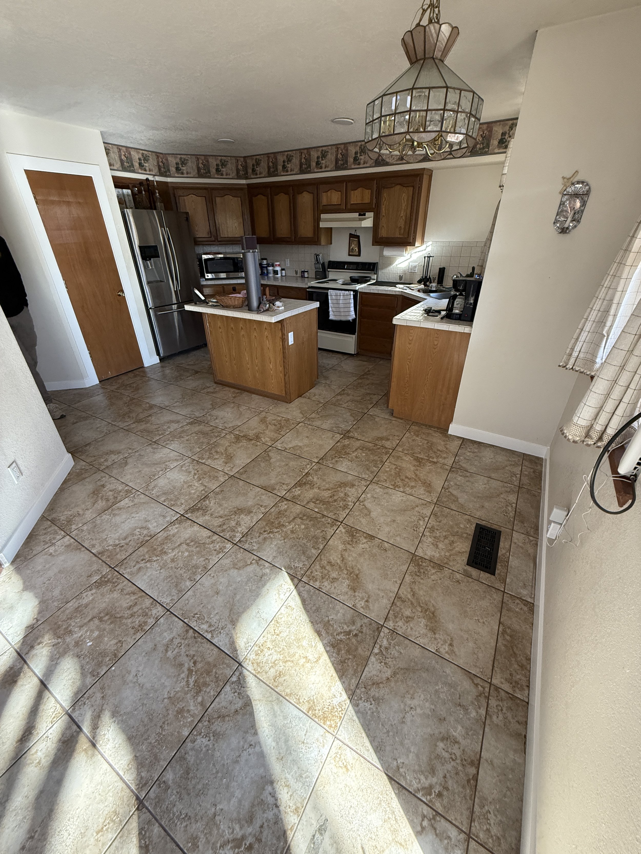 Kitchen with wooden cabinets, tiled floor, and modern appliances including a refrigerator and oven. A small kitchen island is in the center, with a decorative light fixture above. Wallpaper borders the top of the wall.